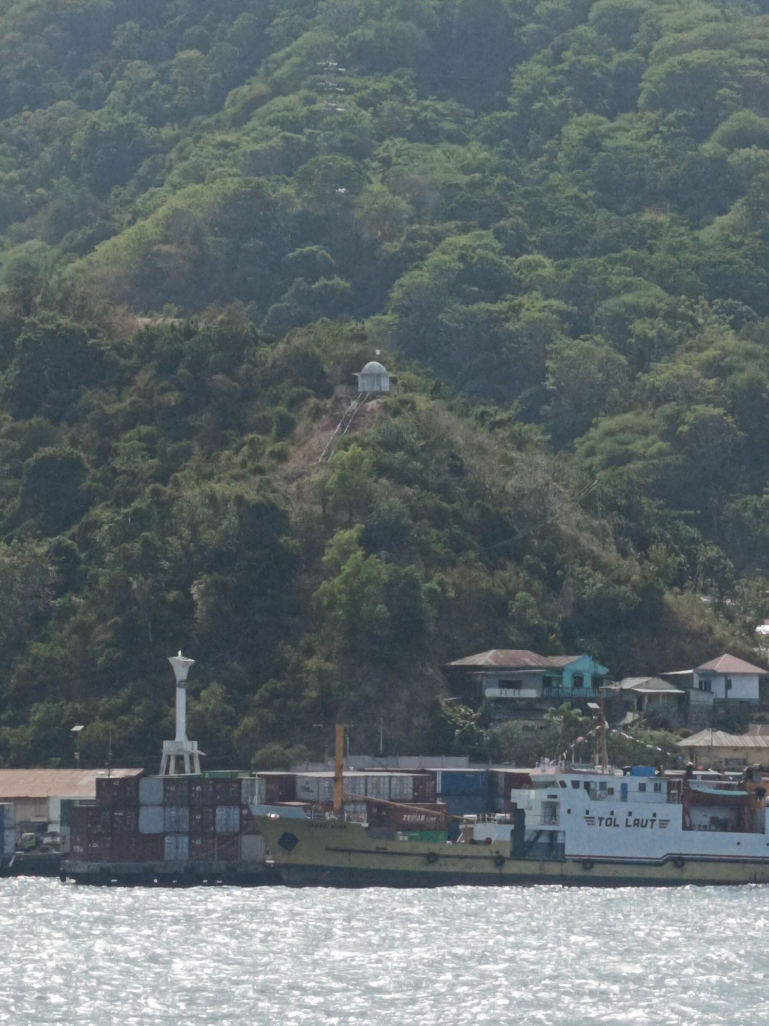 This is a wide-angle, medium-distance photograph of a coastal scene in Gorontalo, Indonesia. In the foreground, the shimmering, sunlit water of the sea occupies the bottom quarter of the frame. A cargo ship, predominantly yellow and white, is docked in the middle ground. The ship bears the name "TOL LAUT" on its side in blue lettering. It is loaded with shipping containers stacked on its deck. To the left of the ship, a small lighthouse-like structure stands on a pier, also surrounded by stacked containers. Behind the dock, a cluster of buildings, including a turquoise-colored house, are visible on the lower slope of a densely forested hill. A pathway leads up the hillside to a small, domed structure at the top. The hill is covered in lush, green vegetation. The sky is a pale, hazy blue, suggesting a bright, possibly humid, day. No people are clearly discernible in the image. The overall impression is of a functional port area situated against a verdant natural landscape.
