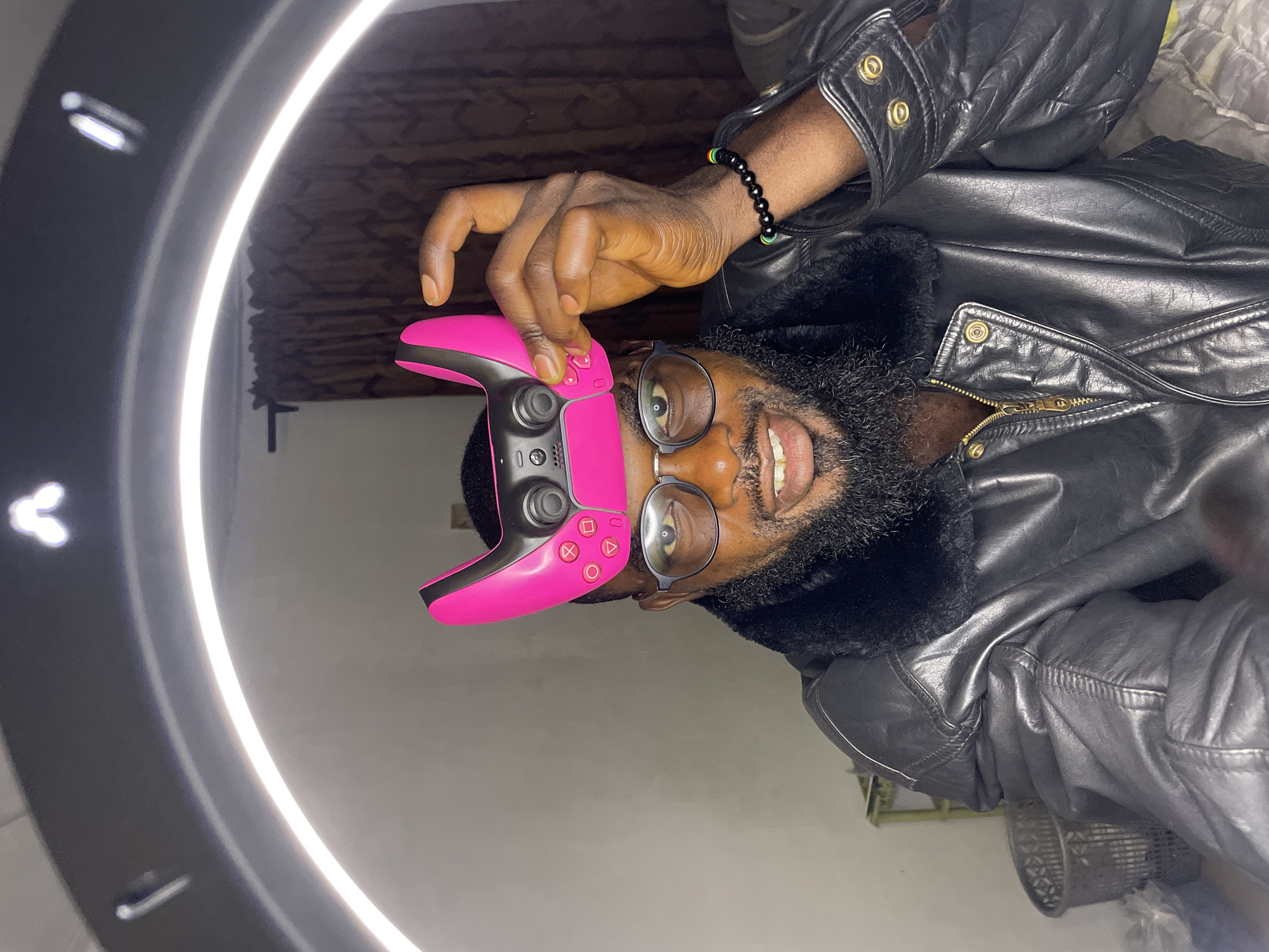 A man is shown with a pink video game controller balanced on his head. He is wearing glasses and a black leather jacket with a fur collar. The background shows a curtain and a ring light is illuminating the scene. The setting is indoors, likely a bedroom, in Jos, Nigeria.