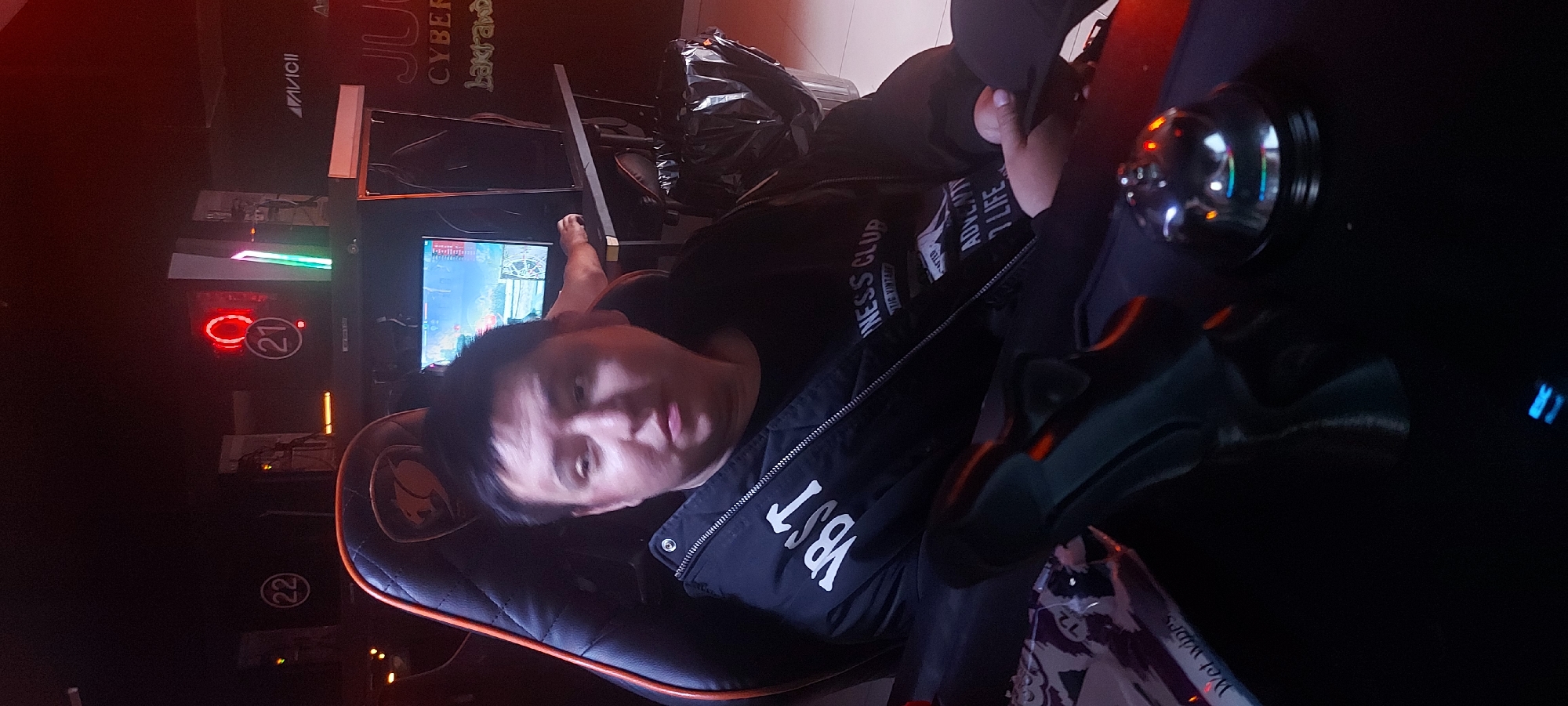 A man is reclining in a black gaming chair in front of a computer monitor. The monitor displays a video game. The room appears to be a gaming center, evidenced by the text "JU CYBER Lakraw" and "AVAVICII" on a panel behind the monitor and the numbers "(21)" and "(22)". The man wears a black shirt and a jacket with "VBST" and the phrase "ADVENTY LIFE" on it. A pack of wet wipes is visible in the lower right of the frame. The location is likely in Tashkent, Uzbekistan, given the text "Lakraw" which might be referring to the gaming center.