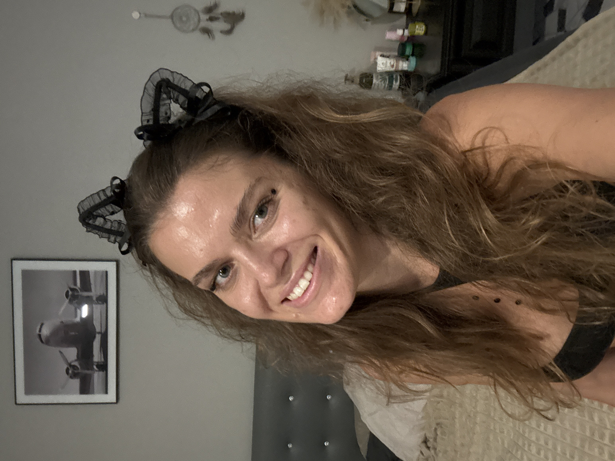 A medium close-up portrait features a woman with long, wavy brown hair and blue eyes, smiling brightly at the camera. She is wearing a black, lacy cat-ear headband. Her head is tilted, and she appears to be in a relaxed, possibly reclining, position. A small dark dot is visible near her left eye, and three small, dark circular marks are on her shoulder or collarbone area, partially obscured by her hair. She wears a dark top.
The setting is an indoor room, likely a bedroom. Behind her, a dark upholstered headboard with decorative light-colored studs is visible, along with a light-colored, textured blanket. The walls are grey. To her left, a framed black and white photograph of an airplane's nose and propellers hangs on the wall. Above her head, a dreamcatcher or similar decorative item is suspended. To her right, shelves or storage units hold various small items, including what appear to be spools of ribbon or thread and bottles.
The woman's broad smile conveys cheerfulness and playfulness. The lighting is soft and ambient, characteristic of an indoor evening setting. The overall scene suggests a casual, personal moment. The location context for this image is Moo 4, Thailand. No visible text is present in the image.