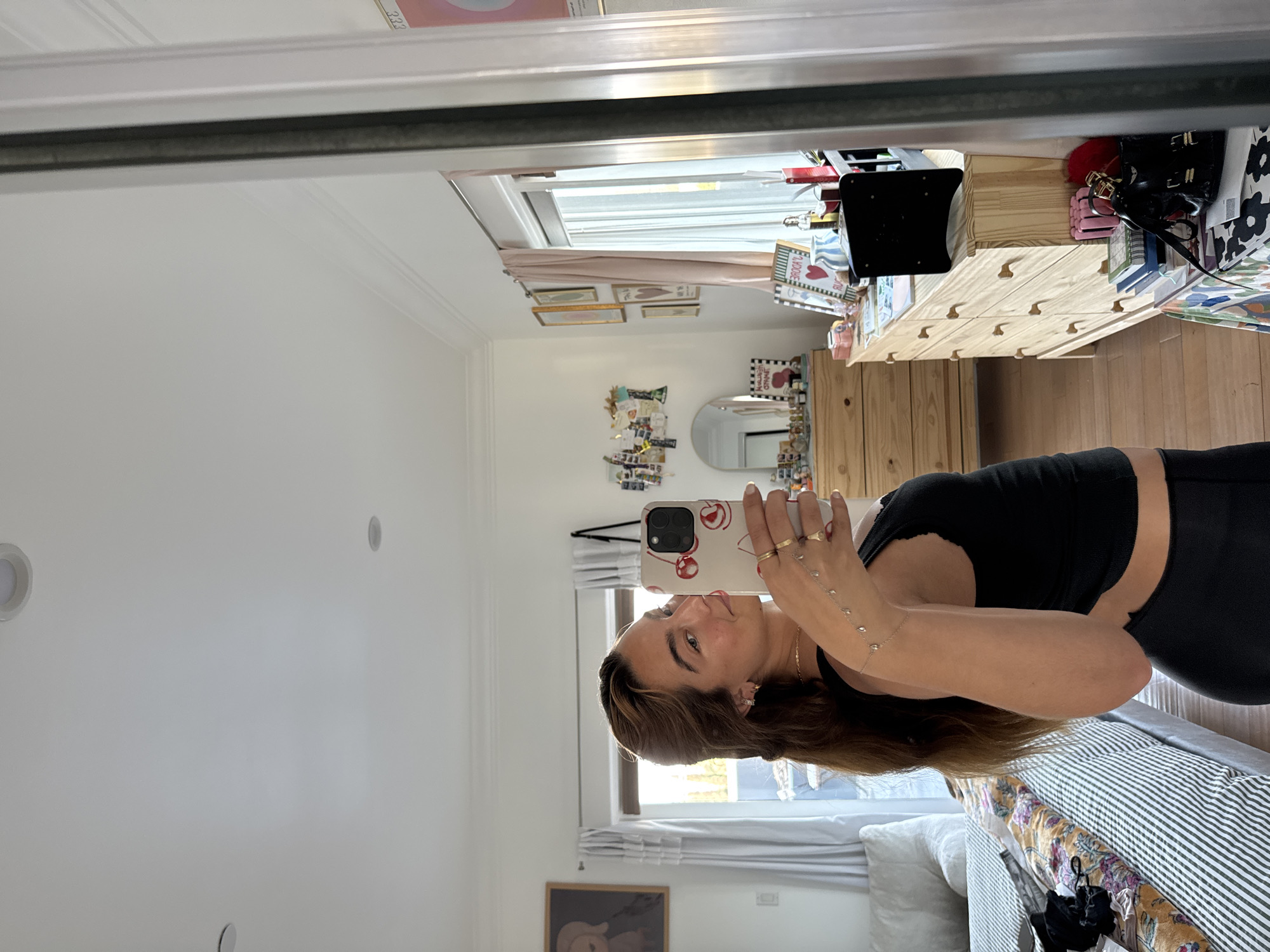 A person in a black tank top is taking a mirror selfie. The room includes a white wall with recessed lighting, a window with white blinds, a white dresser with wooden knobs, framed artwork, and a bed. It is not possible to determine the specific location in Los Angeles from the image.