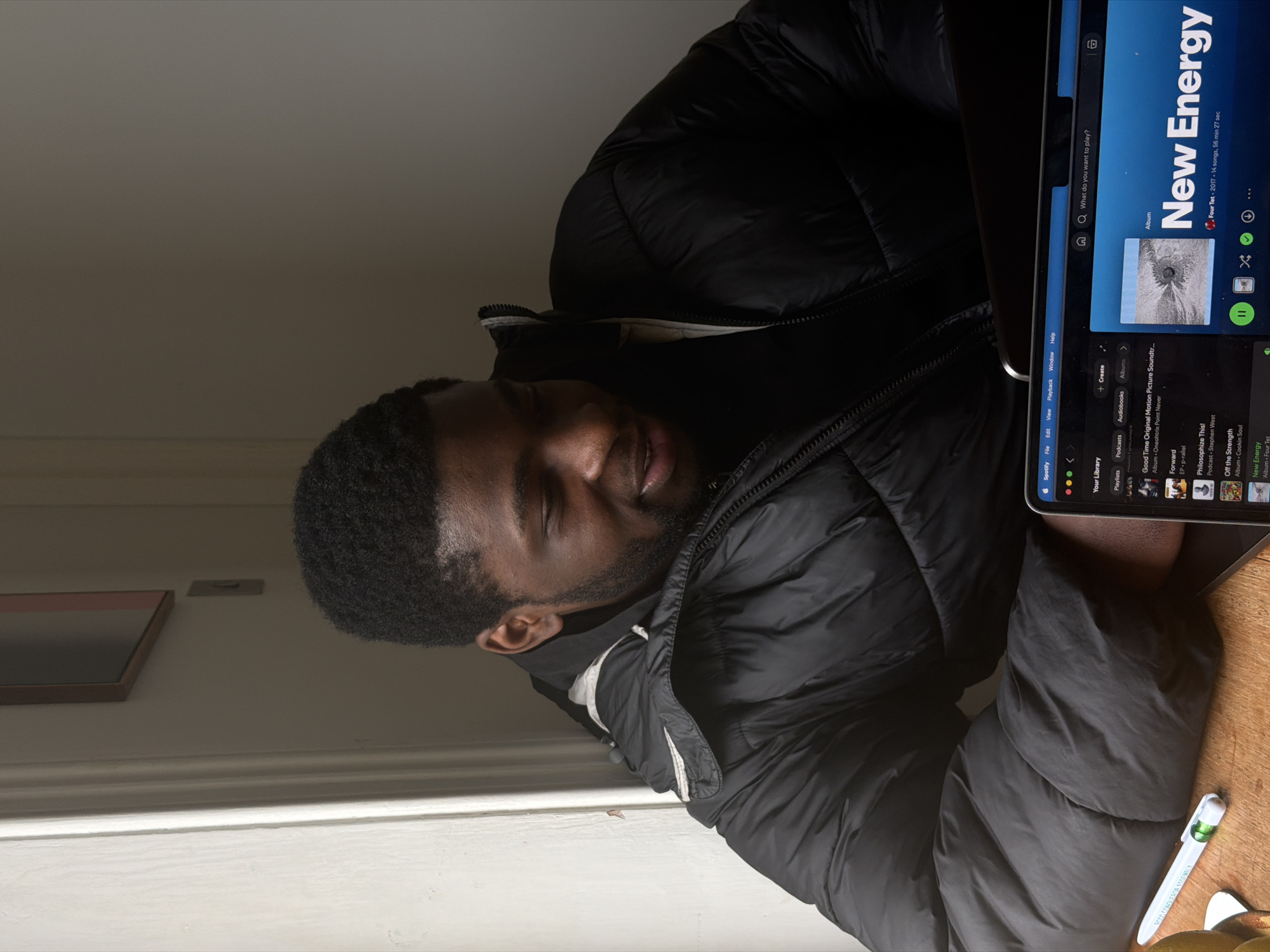 A person is seated at a wooden surface, with a black puffer jacket on. They have short dark hair and dark skin. To their right is a laptop or tablet screen displaying the Spotify application, specifically the album "New Energy" by Four Tet. Next to the device and the person's arm, there's a white pen with green accents. The scene is indoors, near a white wall with a framed picture, and there's ample natural light coming from the side. The precise location is not discernible from the image. The location in London, UK cannot be determined with only the image.