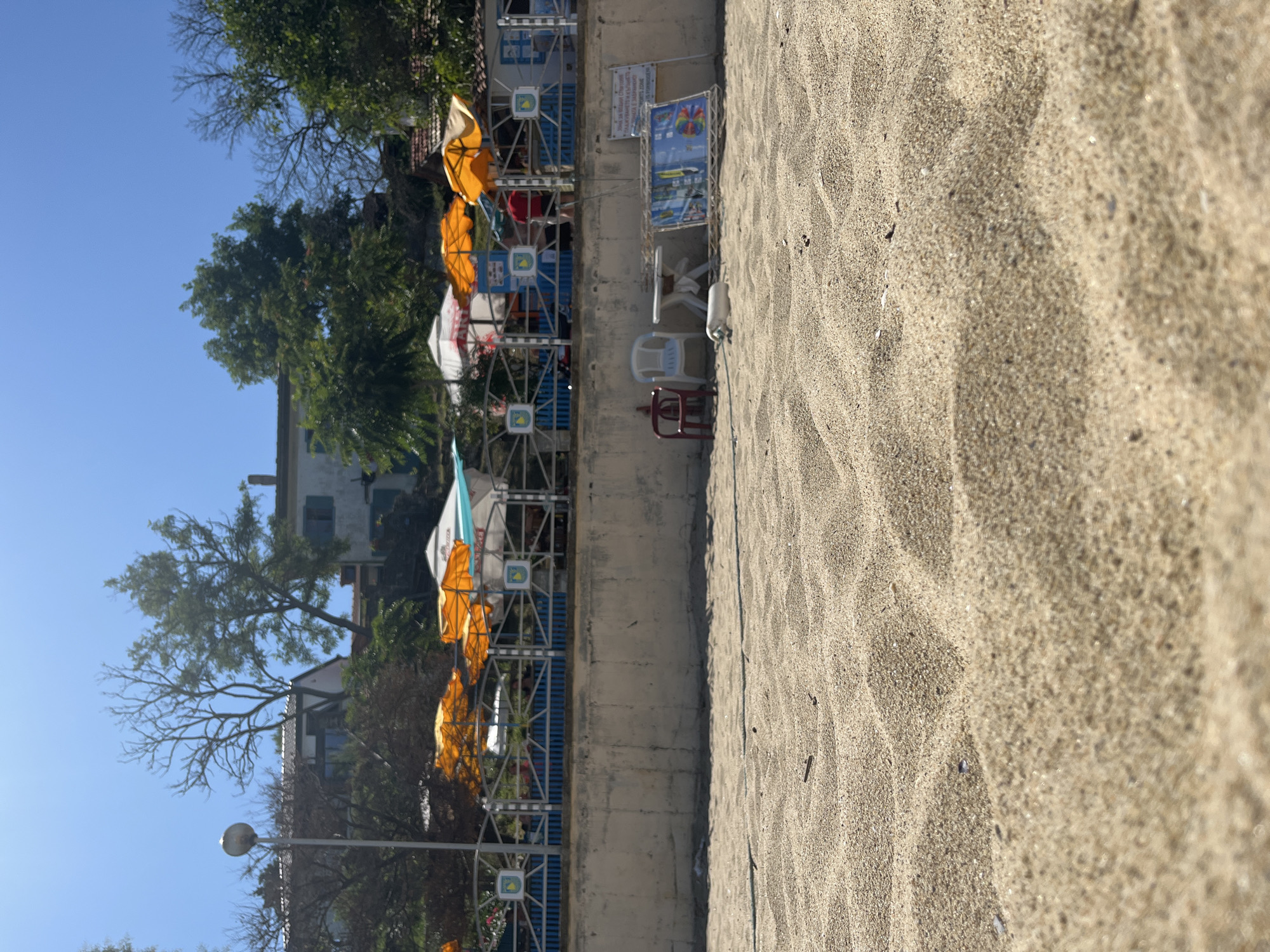 A low-angle shot shows a sandy beach in the foreground, with textured sand dunes stretching across the frame. In the mid-ground, a concrete wall separates the beach from a paved area with various objects. There are two plastic chairs, one red and one white, a small white table, and a large, flat display board with colorful images. A white cylindrical object, possibly a buoy, rests on the sand near the table. Beyond the wall, a fence with blue railings and decorative metalwork encloses a walkway with several open beach umbrellas, mostly orange and white. Behind the umbrellas, a building with a white facade and green shutters is visible, surrounded by trees. A street lamp stands to the left of the scene. In the background, more buildings and trees are seen under a clear blue sky. The location is identified as Obzor, Bulgaria.