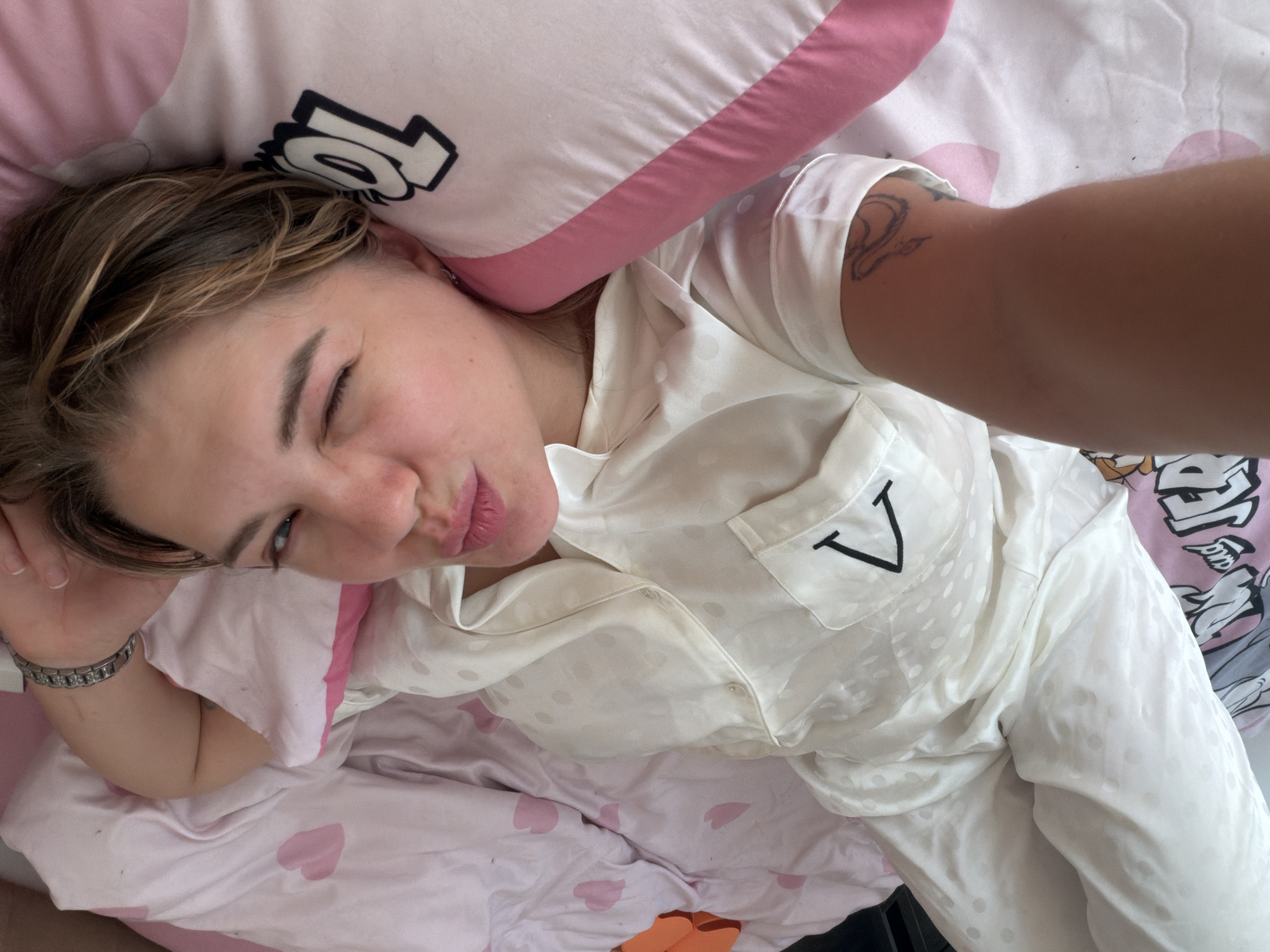 A young woman is lying in bed, presumably in Большая Шильна, Russia, based on geographical context. She is wearing white pajamas with a polka dot pattern and a "V" initial on the chest pocket. She has a bracelet on her left wrist and a tattoo on her right arm. She is winking and pouting at the camera, which she is presumably holding herself to take the picture. She is lying on a bed with a pink and white patterned comforter and a pink and white pillow. The pillow has black stylized letters on it.