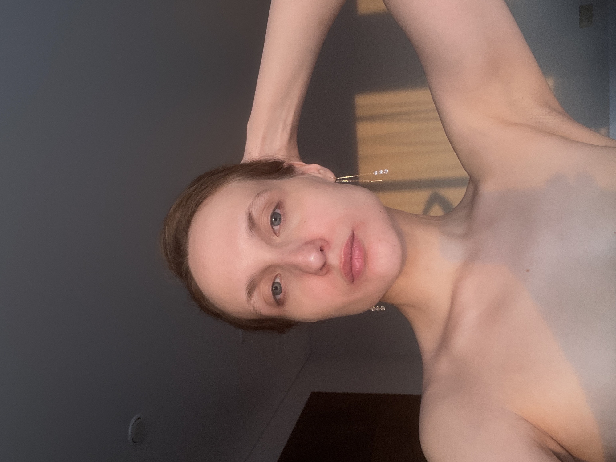 A fair-skinned individual with light brown hair and blue eyes is captured in a close-up, upward-angled portrait. Their head is tilted, and they look directly at the camera with a neutral to slightly serene expression. Parts of their shoulders, upper chest, and arms are visible, suggesting they are shirtless. One arm is raised and bent, with the hand resting behind their head. They wear delicate, gold-toned dangle earrings with small crystals, one featuring three and the other two.
The setting appears to be indoors, against plain dark grey walls. Warm, golden-hour light streams in from an unseen source, likely a window to the right, illuminating portions of the subject's skin and casting distinct light and shadow patterns on the wall and the individual. This lighting creates a warm glow on the skin, contrasting with the darker background. The overall impression is calm and contemplative. No visible text is present in the image. The image is vertically oriented but tilted to the left. The context indicates the location is Podshivalovo, Russia.