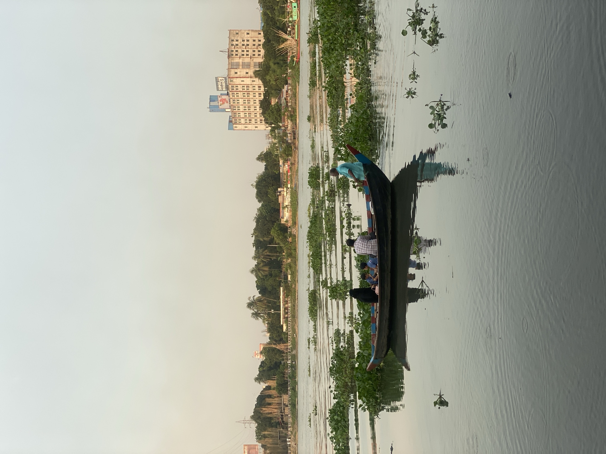 This is a medium shot, captured at eye-level, of a river scene in Dhaka, Bangladesh, under a hazy, pale sky. The foreground is dominated by the calm, gray surface of the river, with subtle ripples emanating from the center, indicating light activity or movement beneath the surface. Scattered patches of water hyacinths float on the water.
In the mid-ground, a long, narrow, dark-colored boat with blue and red trim is prominently featured. The boat contains four individuals: a person in a black headscarf and dark clothing, two individuals appearing to be children in school uniforms, and a man wearing a plaid shirt. Towards the front of the boat, a man in a light blue shirt is visible, seated. The boat is moving through the water, as suggested by the wake behind it and the reflection on the water's surface.
The background depicts the cityscape of Dhaka. A wide riverbank lined with trees and vegetation separates the water from the urban environment. Several buildings are visible, including a tall, multi-story building with a prominent "SUPER BOARD S" sign. Construction activities, including scaffolding and what appear to be makeshift structures, are also evident along the riverbank. Power lines and utility poles are visible in the distance. The overall atmosphere suggests a moderately populated area within the city, likely during daytime given the light.