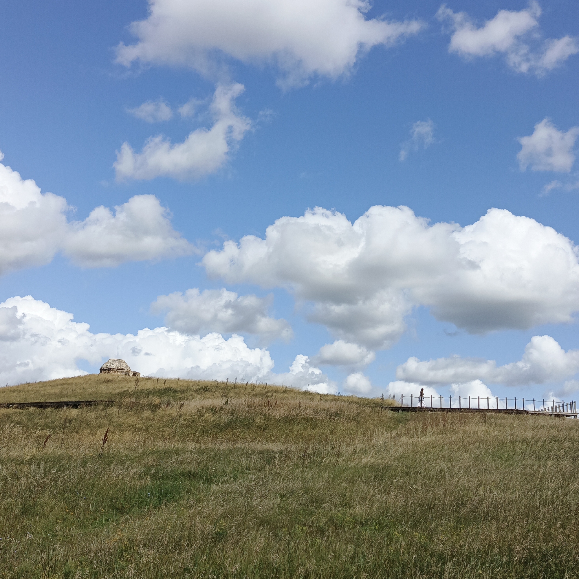 This image shows a landscape with a grassy hill under a blue sky with white clouds. On top of the hill is a small, domed stone structure. In the middle ground, a wooden walkway with a railing extends across the hillside. A lone figure, appearing to be a woman with a bag, walks on the walkway. The foreground is filled with tall, dry grass. The scene is likely located in Verkhnie Termy, Russia, given the architectural style of the structure and the natural environment.