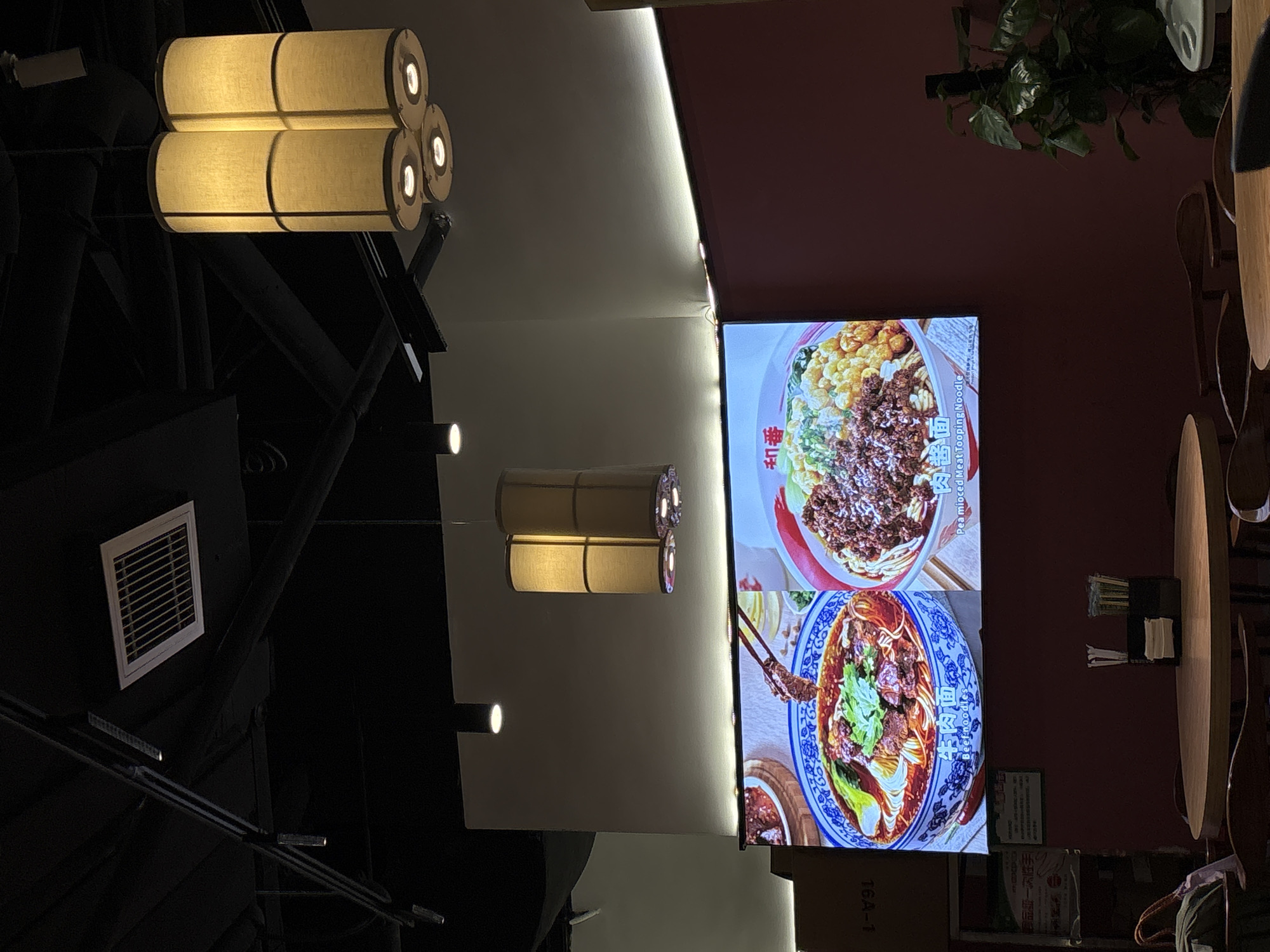 The image depicts the interior of a restaurant in Jianyang City, China. In the foreground, a round wooden table is set with a black napkin holder containing white napkins and chopsticks. The table is surrounded by dark wooden chairs. On the wall behind the table, a large digital screen displays images of two noodle dishes. The left dish is a bowl of beef noodles with broth, and the right dish appears to be a noodle dish with a meat topping and corn. Above the screen, there is a white wall with two hanging cylindrical lamps. The ceiling is black and features exposed industrial elements, including track lighting and an air vent. Further up and to the right, more cylindrical lamps are visible, illuminating the space with a warm glow. A large plant with dark green leaves is situated to the right of the screen, adding a touch of greenery to the scene. The overall ambiance suggests a modern dining establishment.
