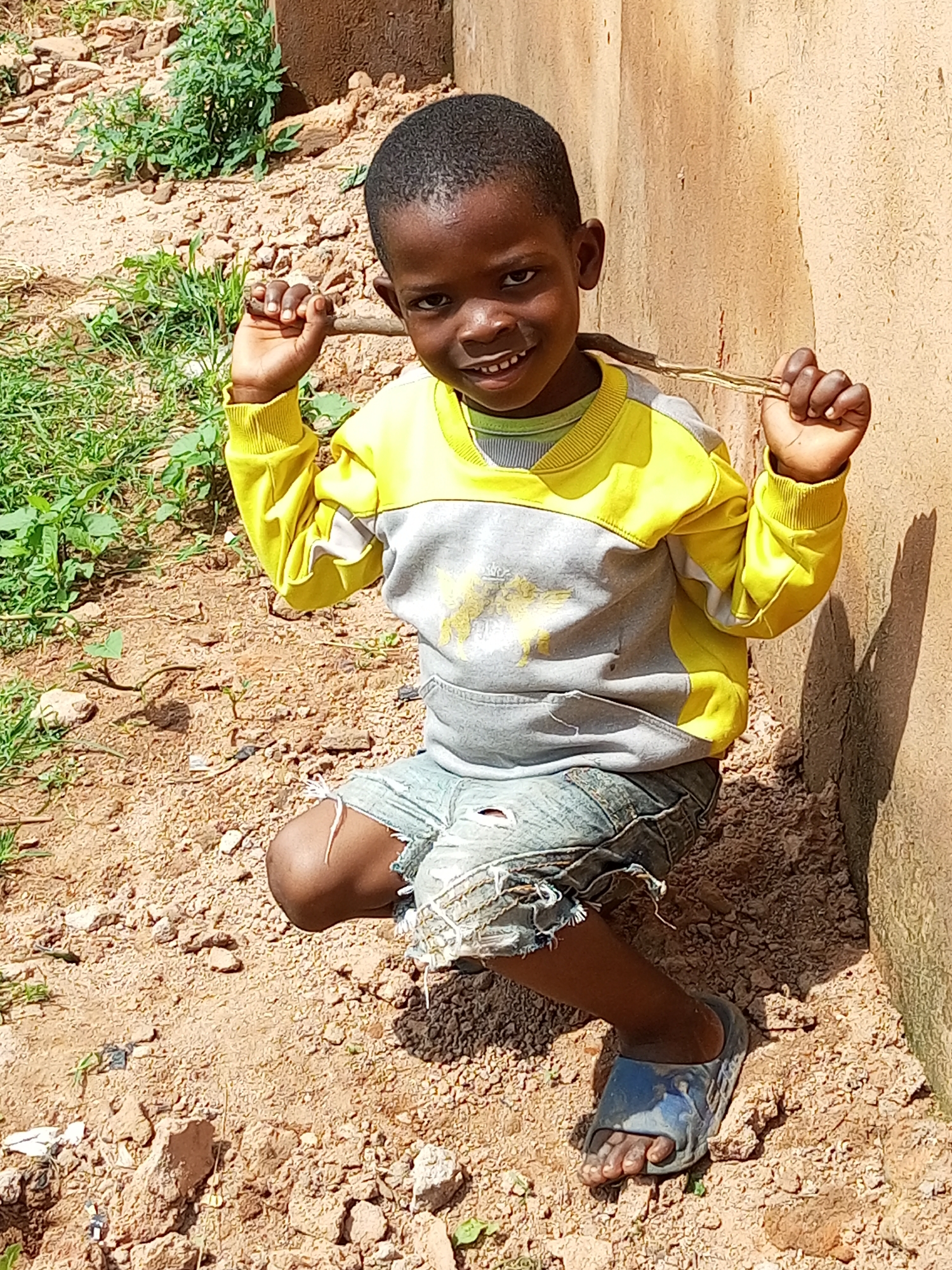 A young Black boy, appearing to be around five to seven years old, is crouched on the dirt ground in front of a textured, tan-colored wall. He is wearing a yellow and gray long-sleeved shirt, denim shorts with rips at the knees, and blue slides on his feet. The boy is holding a thin stick horizontally behind his head with both hands, and he is smiling directly at the camera. The ground around him is sandy with scattered small rocks and sparse green vegetation. The background shows more of the wall and some greenery. The overall impression is of a casual outdoor setting in a rural or semi-rural environment, possibly in Tempure, Nigeria.