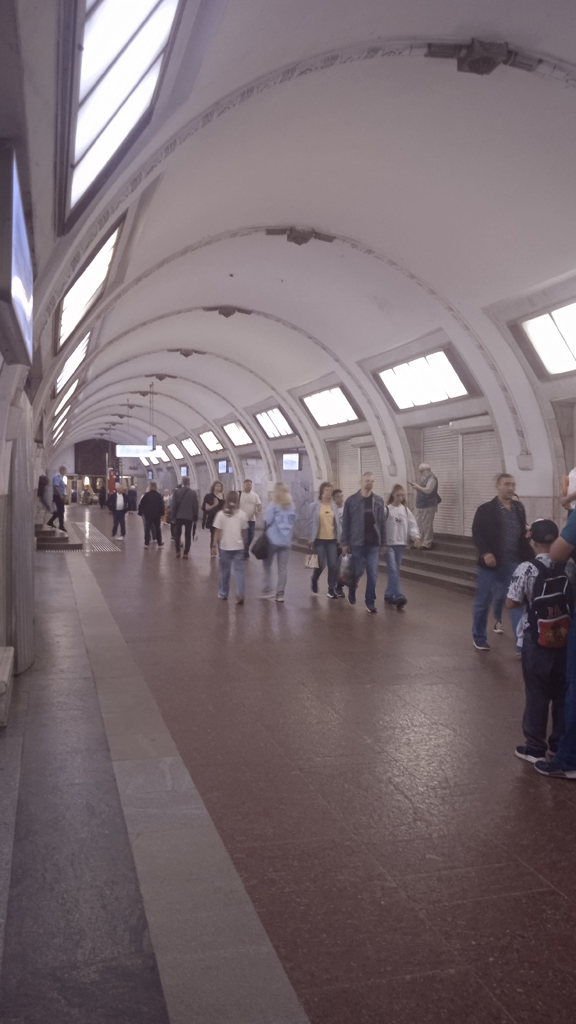 The image depicts a subway station with people walking along a platform. The station has a vaulted ceiling with numerous skylights or illuminated panels. The platform floor is paved with large, reddish-brown tiles, with a lighter colored strip running along the edge. Several individuals are visible, most of whom are walking away from the camera. One man in the foreground on the right is wearing a dark jacket and jeans, with a boy in a checkered shirt beside him. Other people are scattered throughout the platform, some walking in groups and others individually. The overall impression is of a busy, but not overcrowded, public transit space.