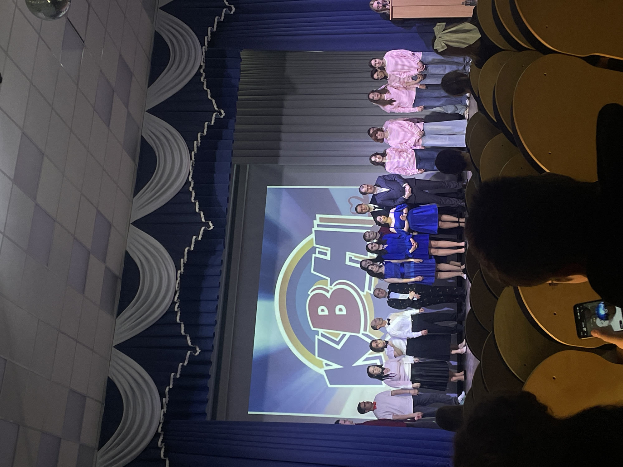 The image shows a group of people on a stage in Novoe Devyatkino, Russia. The stage has a large screen behind the group displaying a logo. The group is arranged in rows, with some wearing pink shirts, others in blue dresses, and some in formal attire. In the foreground, audience members are visible, with one person taking a photo with their phone. The stage is flanked by blue curtains with white decorative trim.