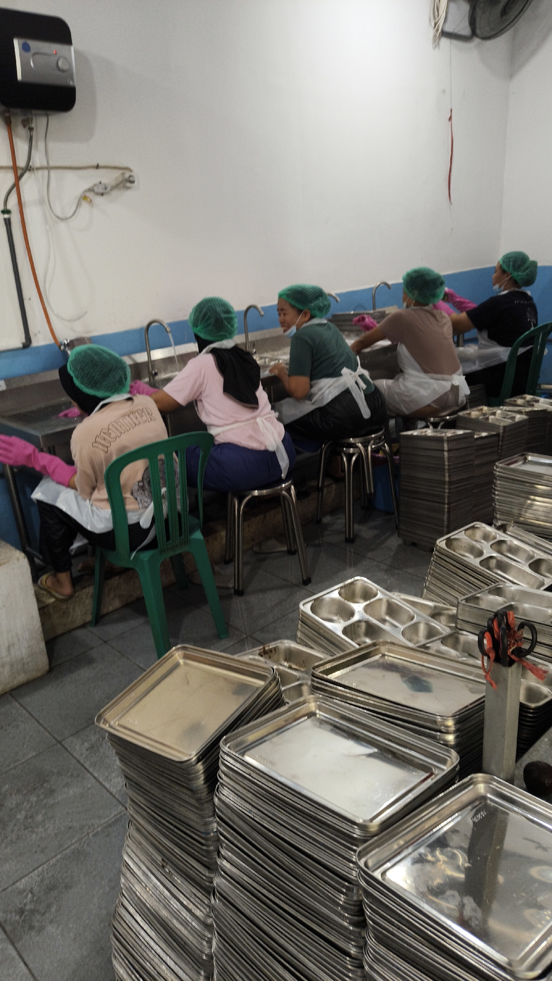 In Sambipondok, Indonesia, a group of individuals wearing green hairnets and aprons are gathered around stainless steel sinks, seemingly engaged in a dishwashing or food preparation task. The environment appears to be a commercial kitchen or service area, with multiple sinks and a large quantity of stacked metal trays and containers visible in the foreground and to the right. The workers are seated on stools and chairs, facing the sinks and working collaboratively. One individual in the center appears to be smiling or laughing, suggesting a relaxed atmosphere. A water heater is mounted on the wall in the background, along with various plumbing fixtures. The overall scene depicts a communal working environment focused on sanitation and food service.