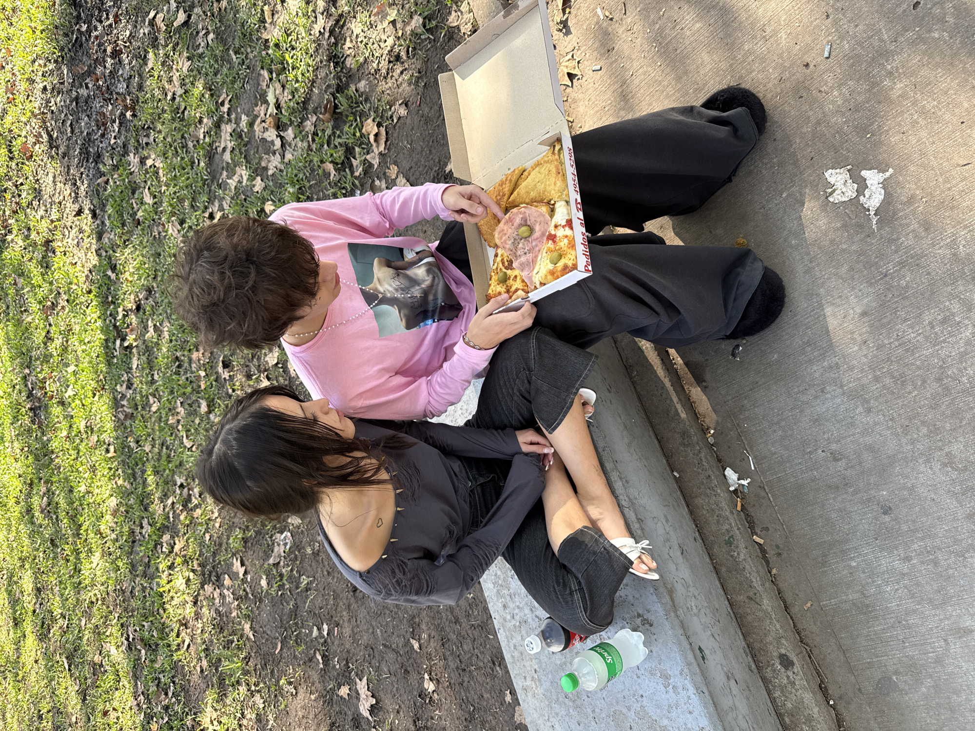 Two people are sitting on a concrete ledge that borders a grassy area with leaves and trees. A person in a pink long-sleeved shirt and black pants with fluffy slippers is holding a pizza box with slices of different kinds of pizza and appears to be selecting a slice. The person is sitting next to another person in a gray top, black jeans, and white sandals. Two plastic soda bottles are on the concrete ledge. A concrete sidewalk is near the ledge. Cigarette butts, trash, and torn paper litter the sidewalk. The pizza box has "Pedidos al 4554 5208" printed on the side. The location is likely in Buenos Aires, Argentina, given the "4554" phone number area code.