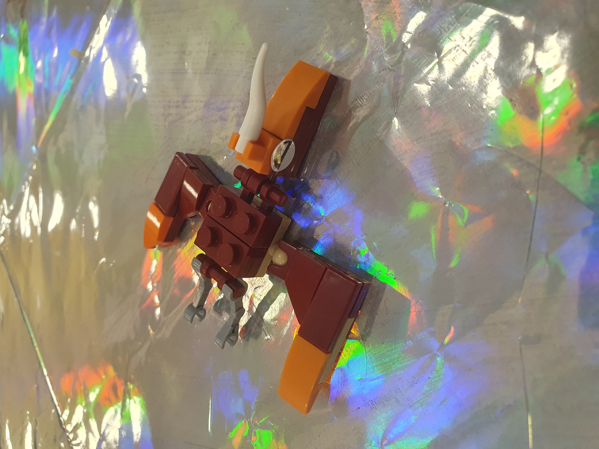 A detailed, forensic, matter-of-fact description of the scene:

The image displays a close-up, angled view of a small, red and orange Lego-built creature lying on a reflective, iridescent surface. The creature resembles a stylized bird or dragon with a predominantly red body constructed from Lego bricks. It has two gray Lego robotic-style appendages extending from its back, possibly representing wings or legs. The front of the creature features an orange Lego piece forming a beak-like structure, topped with a white, curved Lego piece resembling a horn or tusk. A circular, silver Lego piece with a subtle imprint is affixed to the side of the orange beak piece. The overall construction suggests a whimsical, toy-like object.

The background is dominated by the highly reflective, crinkled surface, which exhibits strong holographic effects, reflecting ambient light in a spectrum of colors including green, blue, purple, and orange. These colors are cast as shifting patches and streaks across the surface, creating a vibrant and somewhat abstract environment. The texture of the background appears to be a thin, flexible material, possibly foil or a similar iridescent wrap.

No people are visible in the image. The lighting appears to be ambient, likely from overhead sources, causing reflections and highlights on both the Lego creature and the background surface. The composition focuses on the object, with the background providing a visually dynamic and colorful context.

The location in the city of Leinatal, Germany, is not discernible from the visual information presented in the image. The scene depicts a tabletop or similar flat surface, and the holographic background is a common material used for crafts or decorative purposes, not indicative of a specific geographical location within Leinatal or any other city.