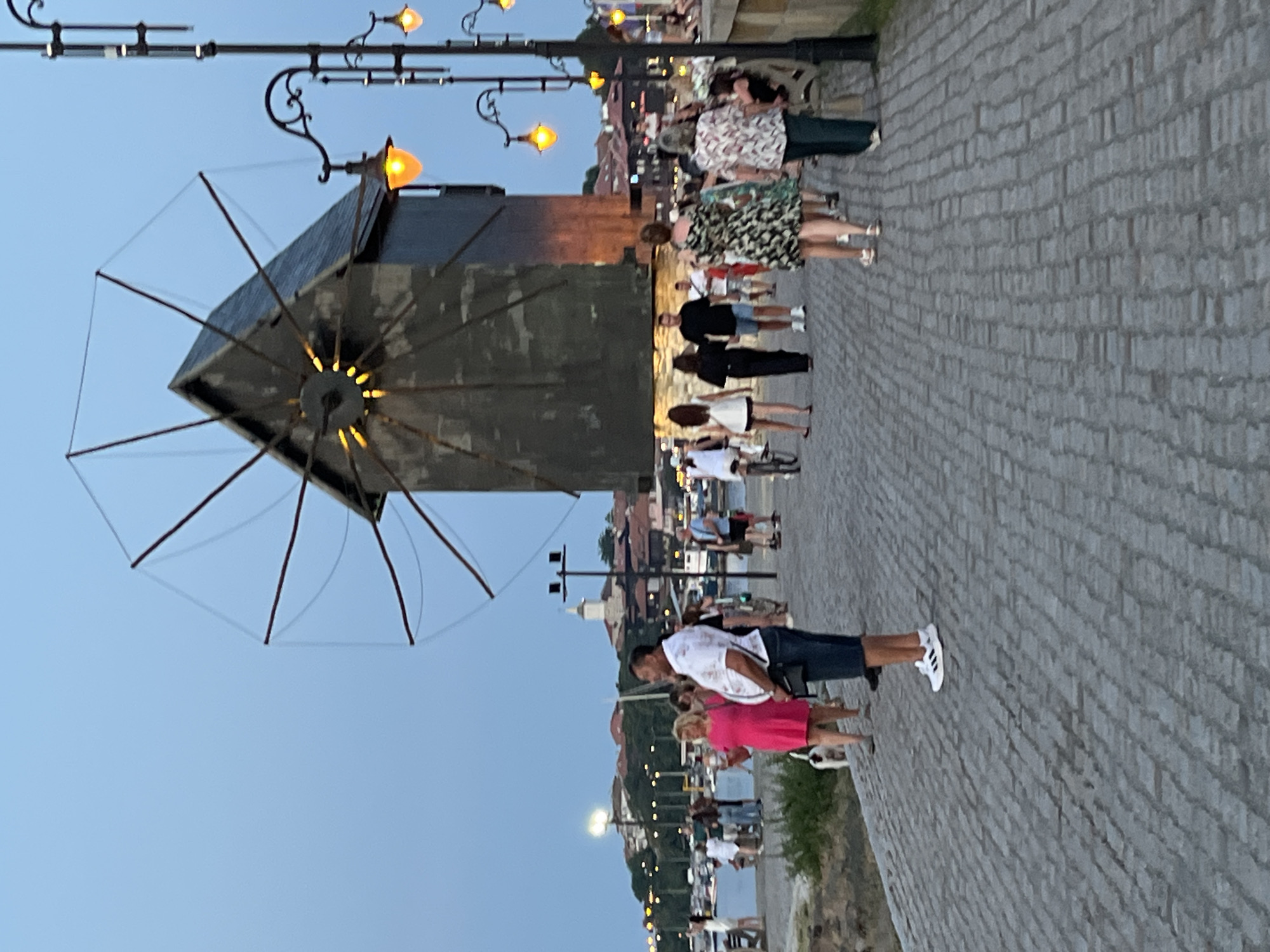 The image depicts a scene in the city of Nesebar, Bulgaria, possibly along its historic promenade or waterfront. A large, dark, weathered structure resembling a windmill with prominent blades dominates the background. The sky is a dusky blue, suggesting either early morning or late evening. In the foreground, a cobblestone walkway is populated by numerous people strolling, walking, and standing. To the right, ornate lampposts with glowing orange lights are visible, illuminating the path. The city's skyline, characterized by terracotta-roofed buildings and a prominent church tower, is visible in the distance across a body of water, likely the Black Sea, where boats are docked. Several individuals are captured in mid-action, walking or talking, with a variety of clothing styles observed, indicating a casual, public gathering. The overall atmosphere is that of a lively tourist area at dusk.
