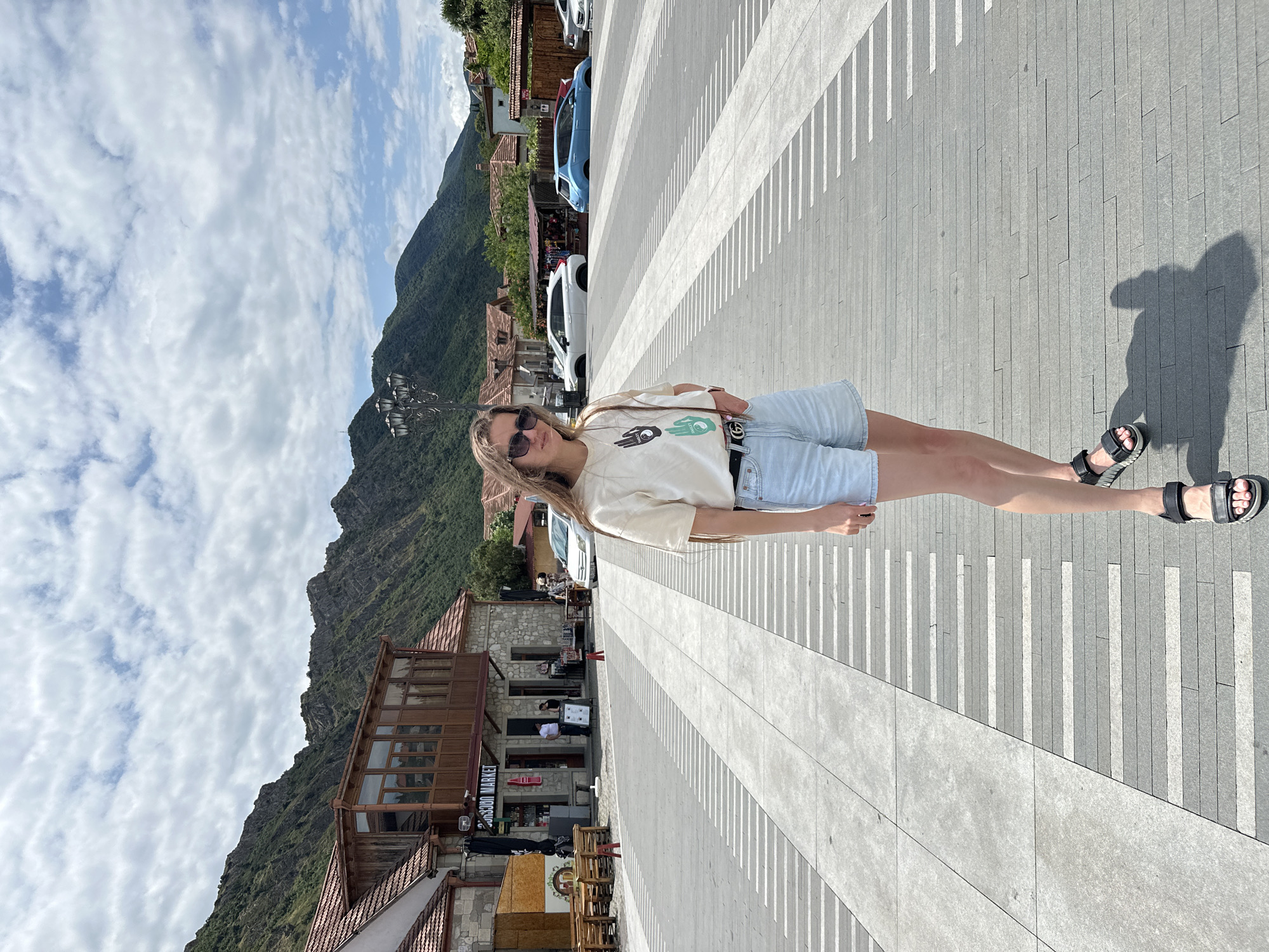 A young woman stands in an open paved square in Mtskheta, Georgia, facing the viewer. She has long, brown hair and wears a cream-colored t-shirt with a graphic print, light blue denim shorts, a black belt with a prominent 'G' buckle, and black sandals. She is also wearing sunglasses and carrying a light brown shoulder bag. The square is paved with large gray stone slabs and a distinct pattern of white and gray rectangular tiles. In the background, there are buildings with traditional red tile roofs and stone facades, suggesting an old town setting. Beyond the buildings, a lush, green mountainside rises steeply under a cloudy blue sky. A few cars are parked in the background, and a street lamp stands to the left of the woman. The overall impression is a bright, sunny day in a historic, picturesque town.