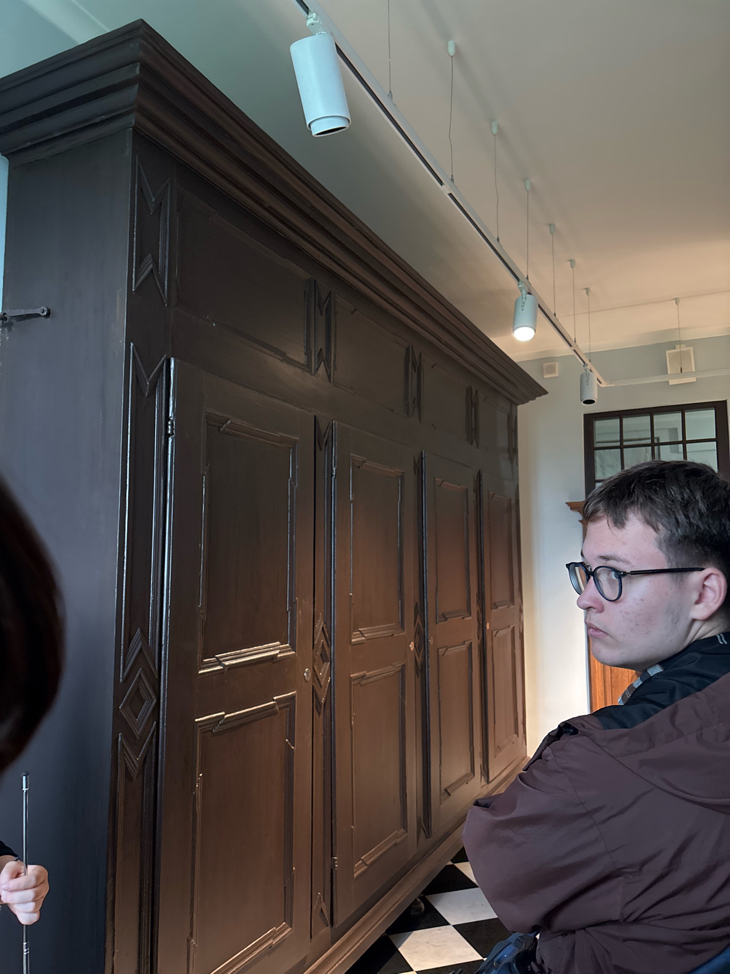 The image depicts a large, dark wooden armoire with intricate carvings on its doors, located in a room with a checkered black and white tiled floor. Several track lights are visible on the ceiling, illuminating the scene. A young person with glasses is positioned to the right of the armoire, looking away from the camera. Another person's arm and hand are visible on the left, holding a slender object, possibly a pointer or a rod. The setting appears to be a museum or historical building, given the ornate furniture and lighting. Based on the style of the armoire and the historical context, this armoire is likely located in the Catherine Palace in Pushkin, a suburb of Saint Petersburg, Russia, possibly within the Amber Room or a similar exhibition space.