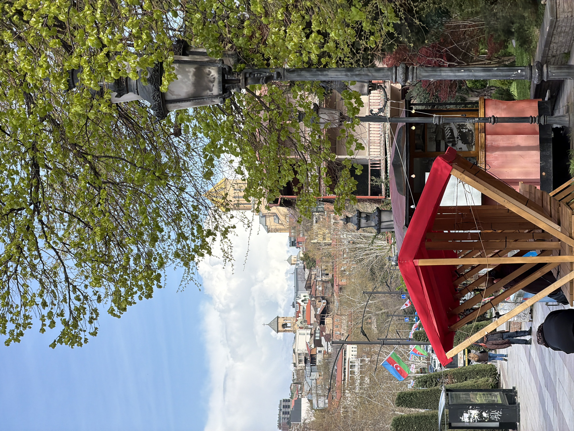 The image shows a street scene in Tbilisi, Georgia. In the foreground, there is a wooden structure with a red awning. A street lamp stands beside it. People are walking along the street. In the background, there are buildings and churches with towers and domes. The sky is blue with white clouds. An Azerbaijani flag is visible.