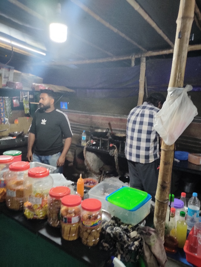 This image captures a bustling night scene at a makeshift food stall or small eatery in Munderi, India. The setting appears to be semi-outdoor, characterized by a dark blue tarp serving as a roof or canopy, supported by rustic bamboo poles visible on the right. Artificial light sources, including a bright overhead bulb and a fluorescent tube, illuminate the area, suggesting it is nighttime.
In the foreground, a counter displays numerous clear plastic jars with red or green lids, filled with various snacks or dry food items. These include what look like savory biscuits, fried snacks, and possibly candies. One jar has a label with "PIRATI" visible. Behind this counter, a man with a beard, wearing a black t-shirt with a white "ADIDAS" logo and blue jeans, stands facing slightly to the left. He appears to be the shopkeeper or an employee, observing the scene or awaiting customers.
Further back, another man in a blue and white checkered shirt and dark pants is seen from behind, attending to a cooking station. This area features at least two gas burners with wok-like pans, indicating food preparation is underway. Various bottles of condiments, beverages, and other supplies are arranged on the counter space around the cooking area and near the bamboo pole on the right. A clear plastic bag is tied to one of the bamboo poles. The overall impression is that of a local street food vendor, busy serving customers after dark.
