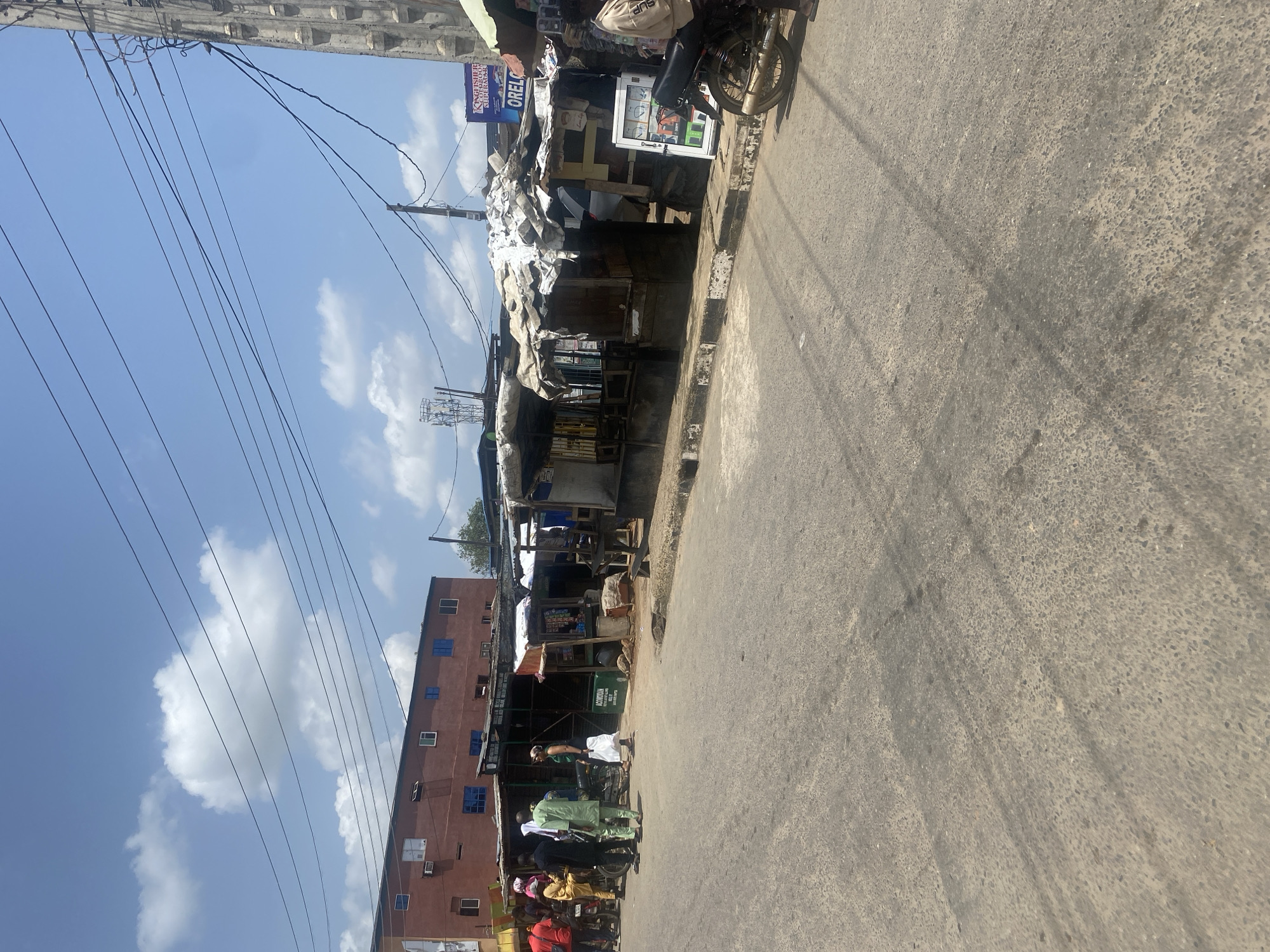 The image shows a street scene in Lagos, Nigeria. Along one side of the street is a row of small, informal shops or stalls under makeshift coverings. Goods are visible in some stalls, and there are people near the stalls, including some on motorcycles. A multi-story building is behind the shops. Overhead are power lines and the sky is blue with some clouds. The street is paved but appears somewhat worn. A sign for "Kingsfisher, Orelc" is visible on one of the stalls.