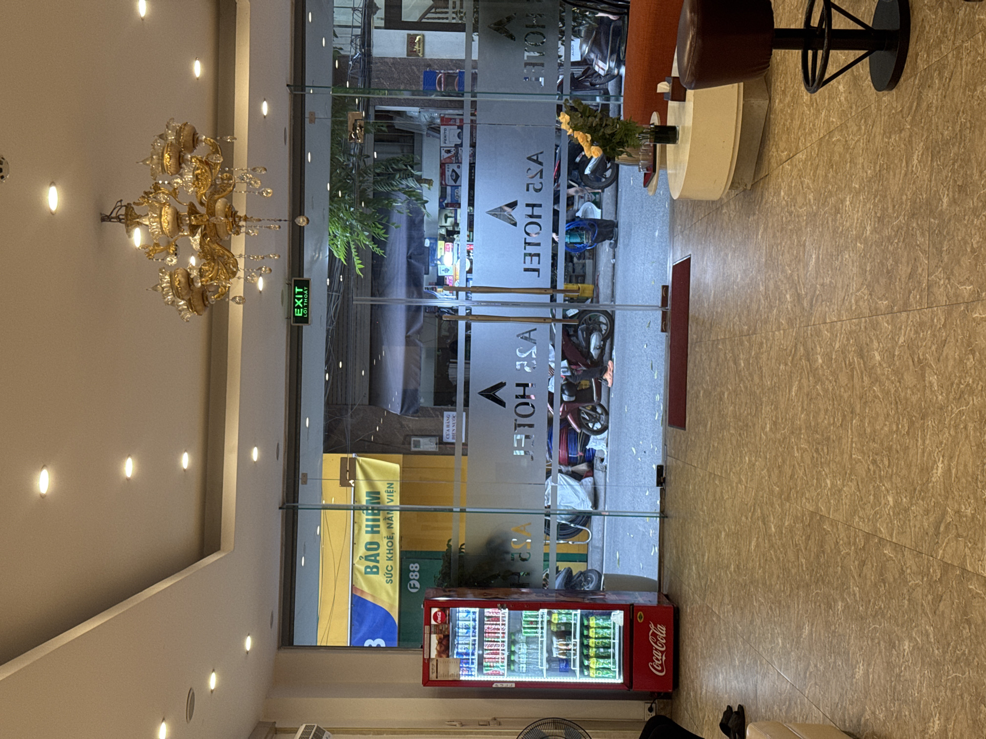 The image shows the lobby of a hotel in Hanoi, Vietnam. A red Coca-Cola vending machine is visible on the left, filled with drinks. Through the glass doors, a street scene is visible with motorcycles and people. The hotel's name, "ASA HOTEL," is frosted onto the glass doors. A chandelier hangs from the ceiling. A table with flowers and a stool are in the foreground.