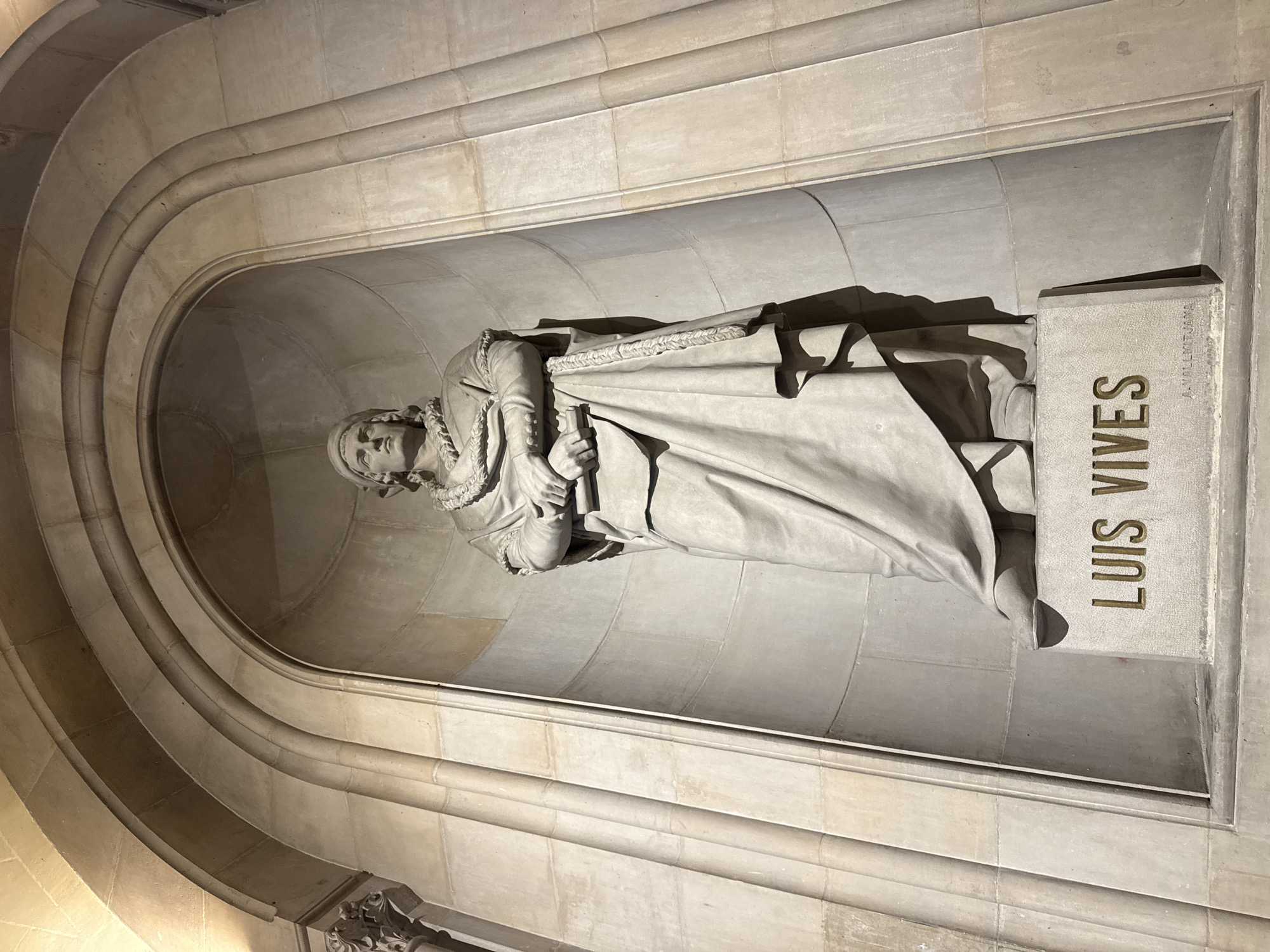 A stone sculpture of Luis Vives (1492-1540) is in an alcove in Barcelona, Spain. He is shown lying down, hands crossed, with a book on his chest. The inscription on the block at his feet reads, "Luis Vives." The interior is made of light-colored stone blocks in an arched design. The name A.VALLMITJANA appears above the inscription.