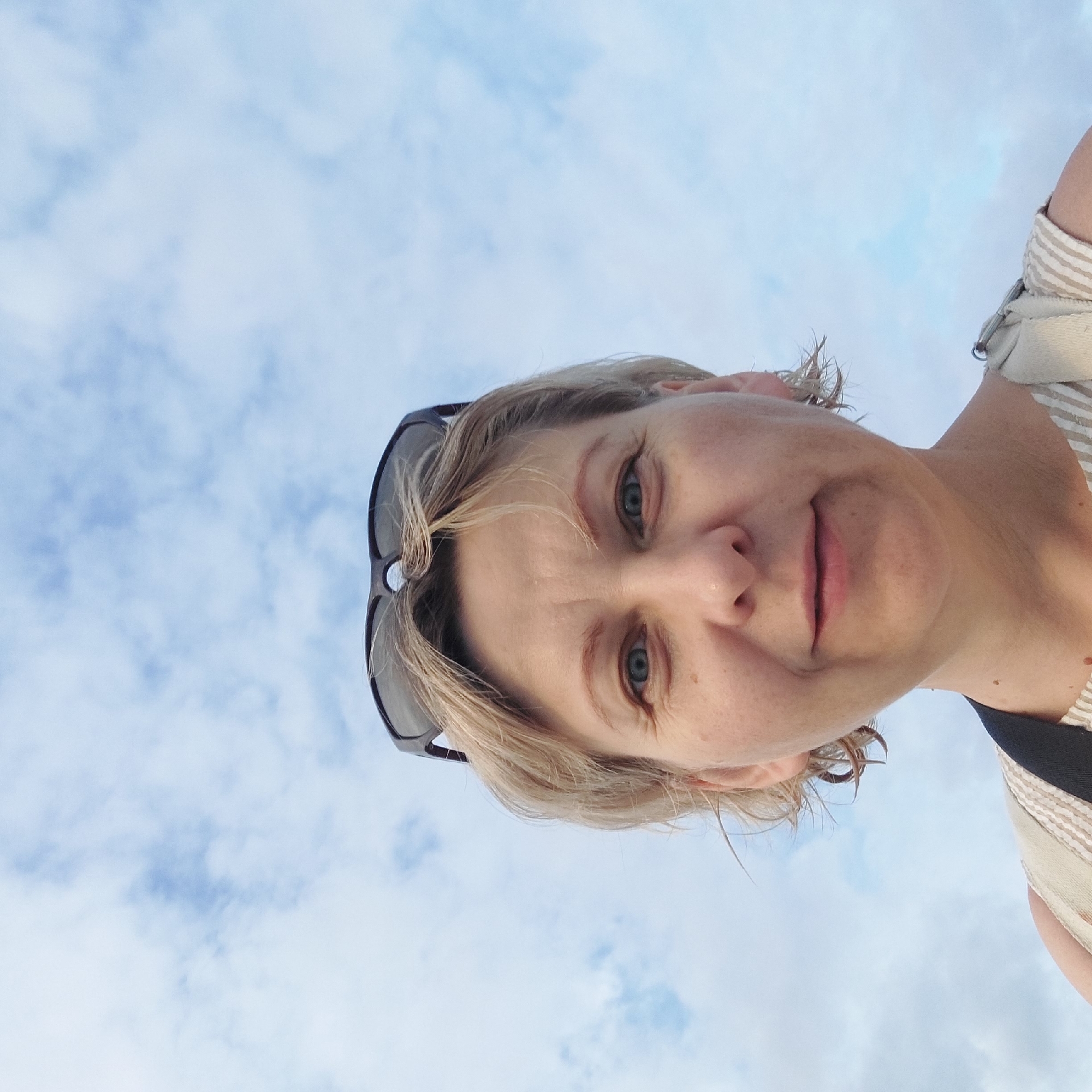 The image is a close-up selfie of a woman with blonde hair and sunglasses perched on her head, against a backdrop of a cloudy sky. The woman is facing the camera, and her face is well-lit. She is wearing a striped top and a dark strap is visible over her shoulder. The image does not contain any information that would allow for the identification of a specific location within the city of Iași, Romania, or any other city.