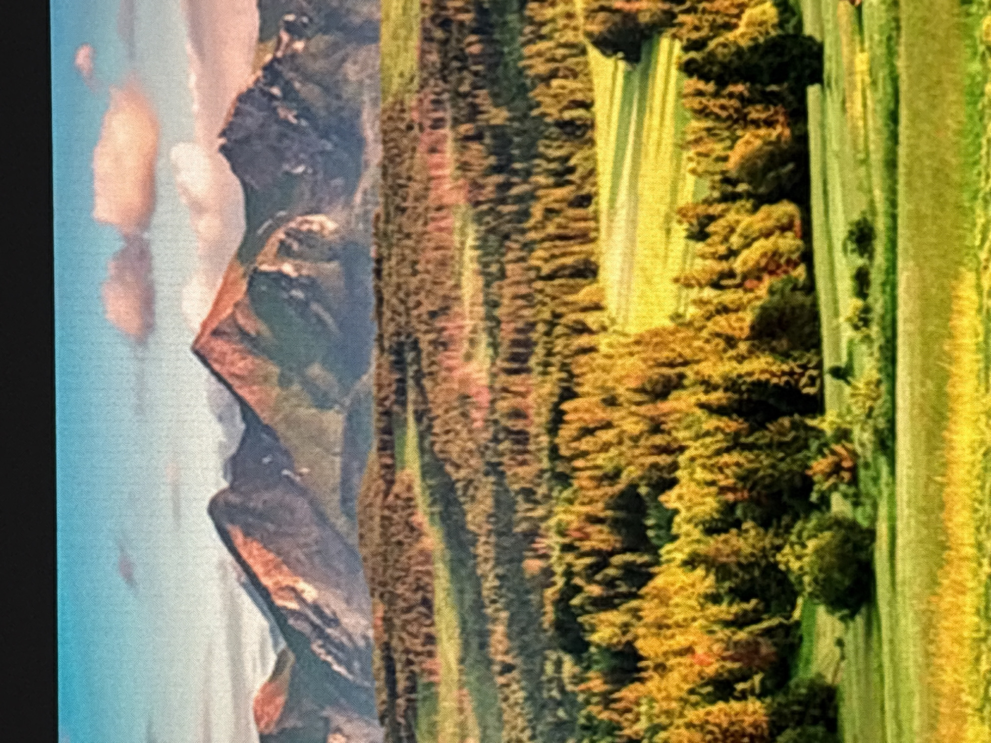 The image shows a landscape scene with mountains in the background, a forest in the middle ground, and fields in the foreground. The sky is blue with some pink clouds. The mountains are rocky and appear to be snow-capped in some areas. The forest is dense and appears to be a mix of evergreen and deciduous trees, with some trees showing autumn colors. The fields are green and appear to be meadows or pastures. The lighting suggests that it is either sunrise or sunset, with the sun casting long shadows. The image does not contain any people, buildings, or any other elements that would indicate a specific location in Sofia, Bulgaria.