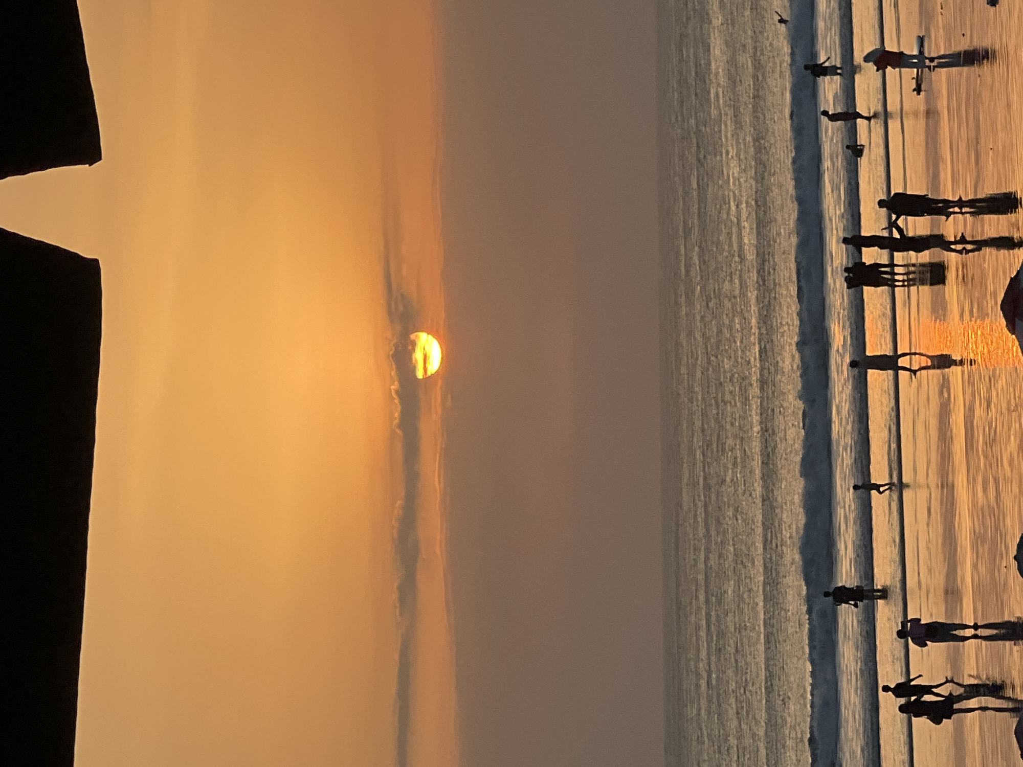 The image captures a sunset on Kuta Beach in Kuta, Indonesia. Silhouetted figures of people are scattered across the wet sand and shallow water, some walking, some standing, and others appearing to take photographs. The ocean in the background displays gentle waves, and the sky is a vibrant orange, with the sun partially obscured by thin clouds. The scene is characteristic of a popular beach destination during golden hour.