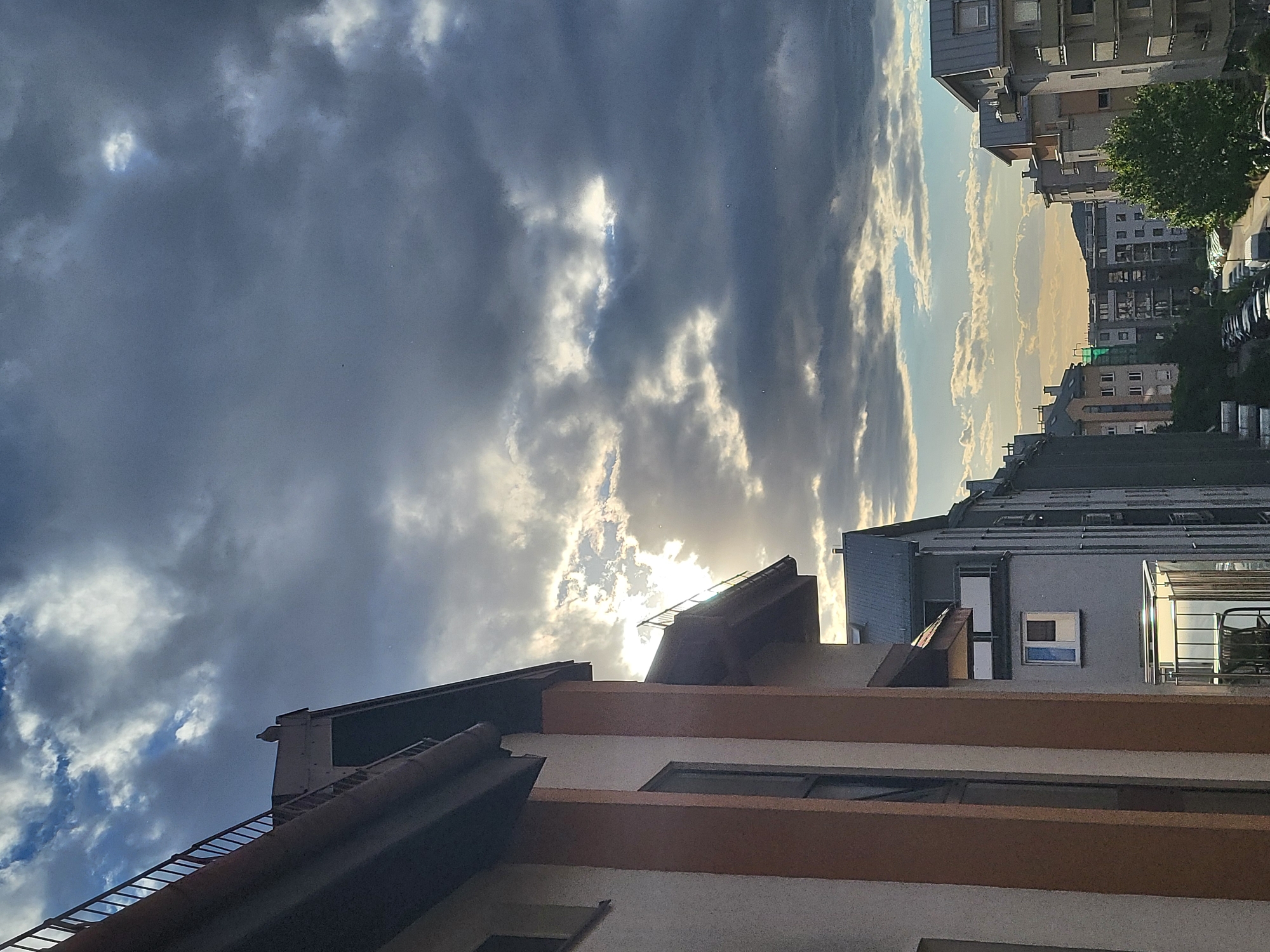 The image depicts a cityscape under a cloudy sky. The foreground is dominated by the roof of a building, featuring dark tiling and orange-painted sections. In the background, taller buildings are visible, receding into the distance along a street. The sky is filled with dark, dramatic clouds, with some lighter patches where the sun is breaking through. The location is Unknown, Unknown. There are no people or interactions visible in the frame.