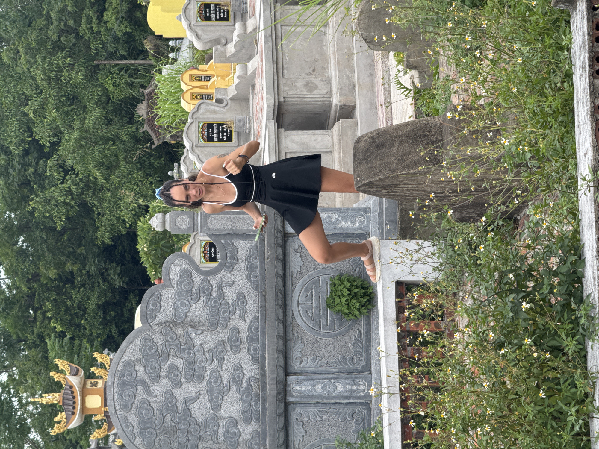 A young woman with braided hair and a blue ribbon is standing on a stone ledge in a cemetery in Hoa Lư Ward, Vietnam. She is wearing a black athletic dress and pink sandals. Her right hand is raised, and she is holding a green smartphone in her left hand. She is looking at the camera with a neutral expression. The cemetery contains many stone tombstones and mausoleums, some adorned with intricate carvings and golden embellishments. Lush green trees and foliage surround the area, with some small white flowers growing in the foreground. The lighting suggests it is daytime with overcast conditions.