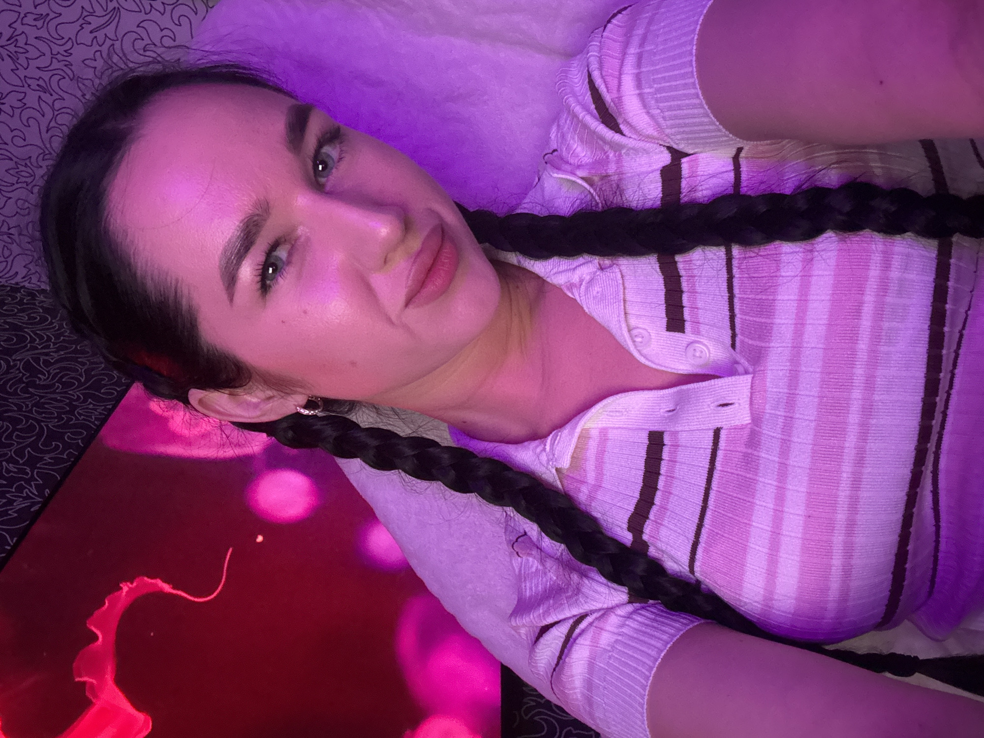 A young woman with dark hair braided into two pigtails is looking at the camera. She is wearing a pink and white striped polo shirt. The background is dark with a pink neon light on the left side and a patterned wallpaper on the top right. The image is illuminated by purple lighting. The location is not specified in the image, therefore the city of Novosibirsk, Russia cannot be identified.