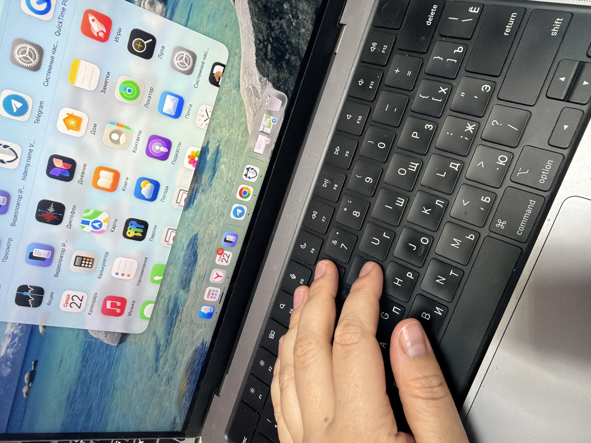 The image captures a close-up, angled view of a person's right hand interacting with a laptop keyboard, while a separate device screen is displayed on the laptop's monitor.
The primary subjects are a human hand, a laptop computer, and a smaller, distinct screen, likely from an Apple iPad or iPhone, positioned over the laptop's display. The laptop keyboard features both Latin and Cyrillic characters, suggesting it is set up for use in a country like Russia, consistent with the provided location context of Chaikovsky, Russia. The person's right hand rests on the keyboard, with fingers positioned over the 'Г' (G), 'П' (P), 'И' (I), and 'Т' (T) keys, indicating active use or preparation for typing.
The laptop's display shows a background image of a tranquil seascape or lake with distant land. Overlaid on this is the separate screen, which appears to be physically resting on the laptop's display rather than being a mirrored software interface. This screen displays a grid of application icons, characteristic of Apple's iOS or iPadOS operating system, many of which have Russian names. Visible app names include "Telegram," "Игры" (Games), "Заметки" (Notes), "Локатор" (Locator), "Почта" (Mail), "Дом" (Home), "Контакты" (Contacts), "Подкасты" (Podcasts), "Дневник" (Journal/Diary), "Книги" (Books), "Погода" (Weather), "Пароли" (Passwords), "Калькулятор" (Calculator), "Календарь" (Calendar) showing "Среда 22" (Wednesday 22), "Музыка" (Music), "Напоминания" (Reminders), and "Системные нас..." (System Settings/Preferences). The laptop's dock, partially visible at the bottom of its screen, contains icons such as Safari, Messages, Mail, Maps, App Store, Settings, and a Trash Can.
Notable details include the dual-language keyboard, the specific Russian app names, and the date "Wednesday 22" on the calendar app, possibly indicating the day the photo was taken. The overall scene suggests a contemporary digital workspace.