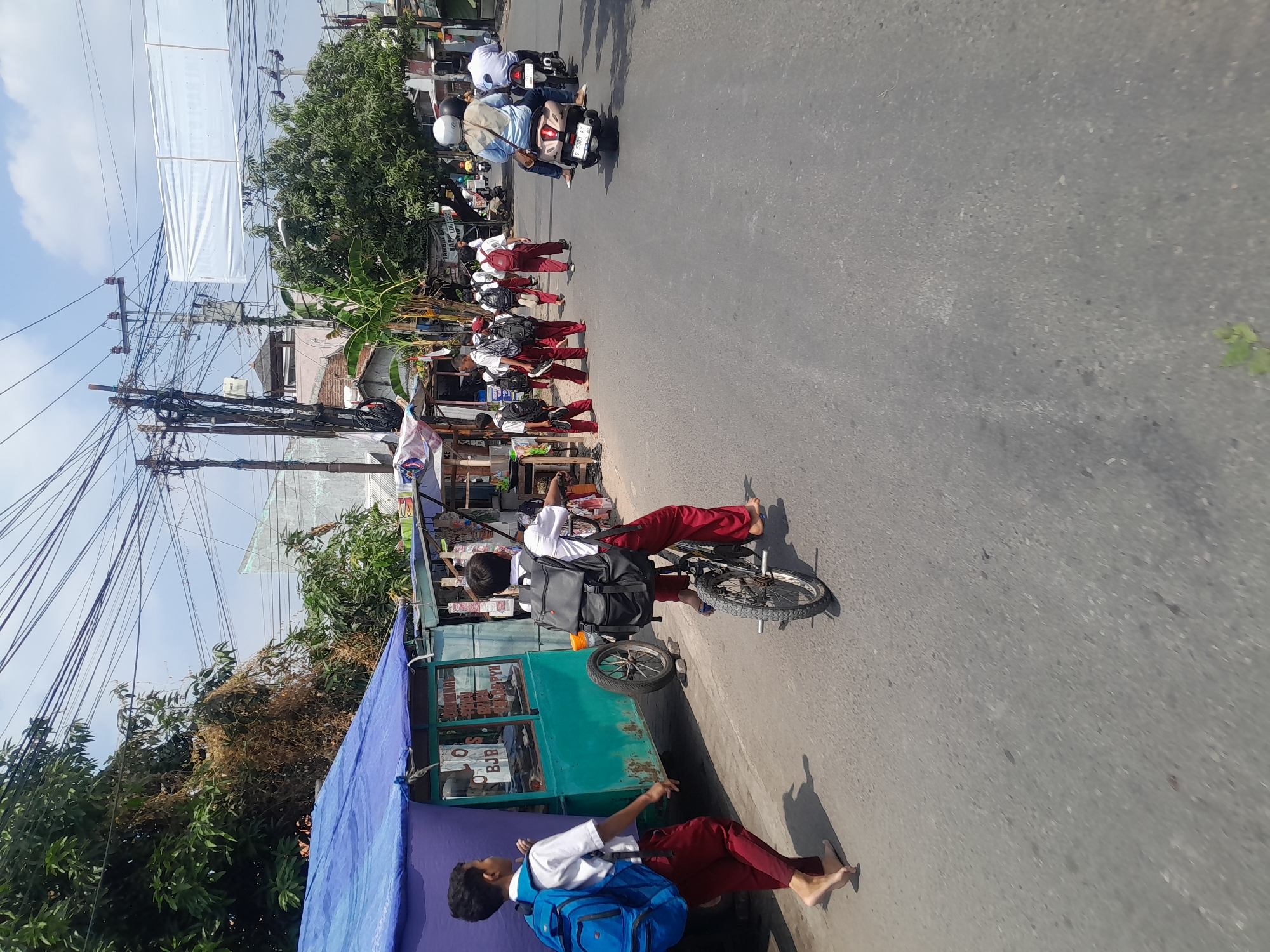 The image depicts a street scene in Tegal, Indonesia, on a sunny day. In the foreground, two young schoolboys in white shirts and red pants are visible. One boy, on the left, walks barefoot with a blue backpack. The other boy, on the right, rides a bicycle and also wears a backpack. A green food cart with a blue tarp roof is parked on the left side of the street. Further down the road, a group of children in similar school uniforms are walking, some carrying backpacks. Several motorbikes are also present, with people riding on them. The street is paved with asphalt, and there are trees and buildings lining the sides. Power lines are strung across the sky. The overall impression is of a typical urban scene in Indonesia, possibly on a school day.