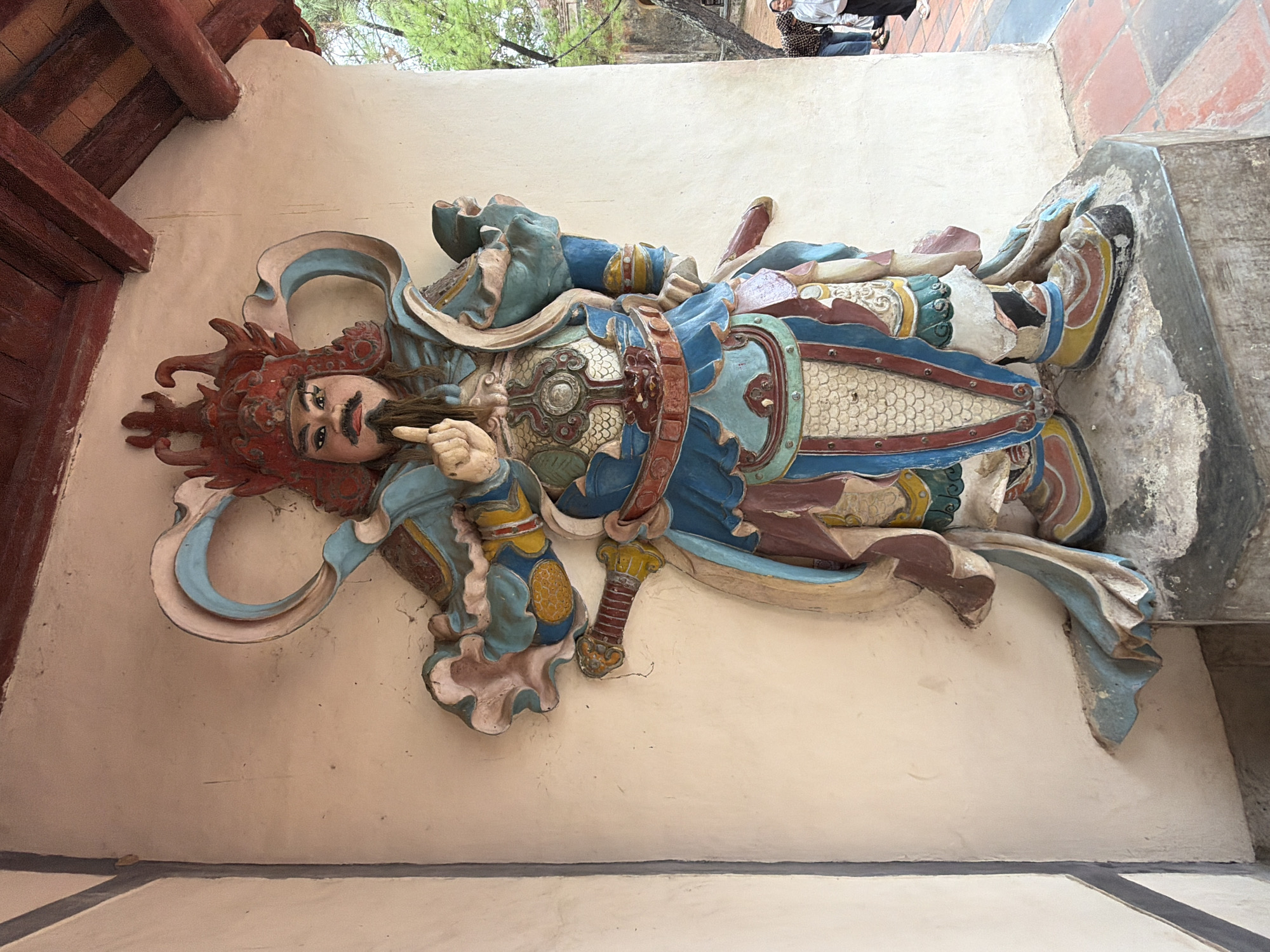 The image shows a full-body statue of a warrior, likely a historical or mythical figure, mounted on a pedestal against a light-colored wall. The statue is richly colored with blues, reds, yellows, and browns, depicting intricate armor and a headdress. The warrior has a stern expression, a mustache, and a beard, and is pointing a finger upwards with his left hand. His right hand is holding a sword, the hilt of which is visible. The statue appears to be made of plaster or concrete and is somewhat weathered. Behind the statue, the architecture suggests an outdoor temple or historical site, with a wooden-beamed roof and tiled flooring visible. In the background, two individuals, a woman and a man, are partially visible, with the woman appearing to be taking a photograph with her phone. The context provided places this scene in Kim Long Ward, Vietnam, suggesting it is a cultural or religious landmark.