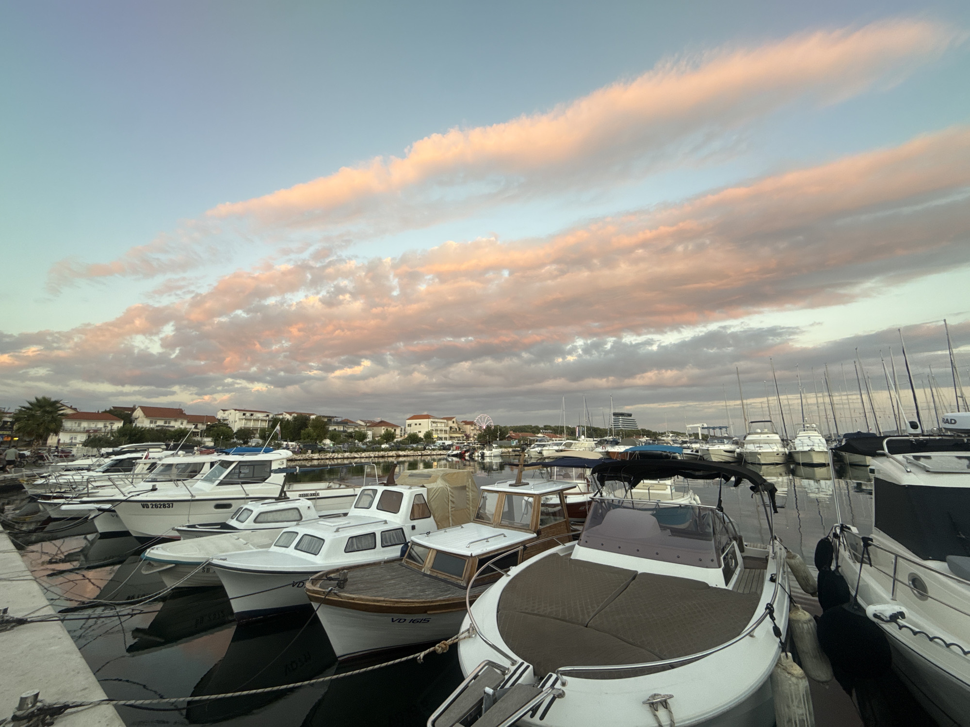 A marina in the city of Vodice, Croatia, is captured at dusk. The sky is painted with soft, pastel hues of pink, orange, and blue, illuminated by the setting sun. Several boats of various sizes are docked along the pier, their white hulls reflecting in the calm water. The boats range from small motorboats with enclosed cabins to larger vessels. In the background, the city skyline is visible, with buildings and palm trees lining the shore. A Ferris wheel stands out against the horizon, suggesting a nearby amusement area. The overall atmosphere is serene and picturesque, depicting a peaceful evening in a coastal town. There are no people visible in the image.