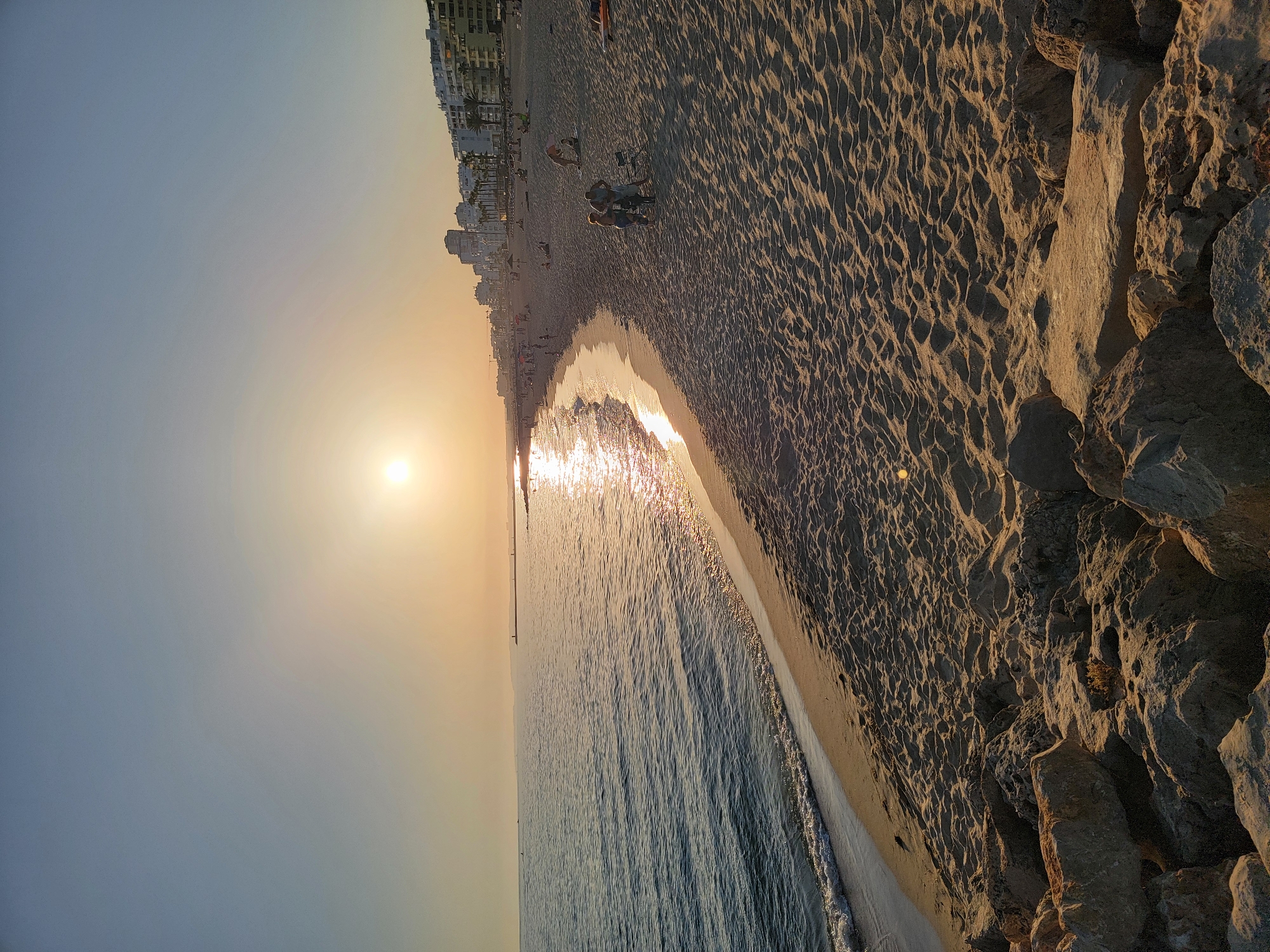 The image depicts a serene beach scene at sunset in Quarteira, Portugal. The sky is a gradient of pale blue and soft orange, with the sun low on the horizon, casting a warm glow across the water and sand. Gentle waves lap the shore, their crests reflecting the sunlight. The beach itself is wide and sandy, with ripples and footprints visible on the surface, indicating recent activity.
In the distance, a line of buildings, characteristic of a coastal resort town, stretches along the coastline, with palm trees interspersed among them. Several people are scattered across the beach, some walking, some sitting, and a few groups interacting. Two individuals are the most prominent subjects in the mid-ground, standing near a foldable chair and a bag. They appear to be engaged in a conversation. Further down the beach, other figures are visible, some sunbathing, others strolling.
The foreground of the image is dominated by a collection of large, weathered rocks, likely part of a coastal defense structure or natural formation, adding a textured and rugged element to the otherwise smooth expanse of sand and sea. The overall atmosphere is peaceful and idyllic, capturing a typical evening at a popular Portuguese beach.