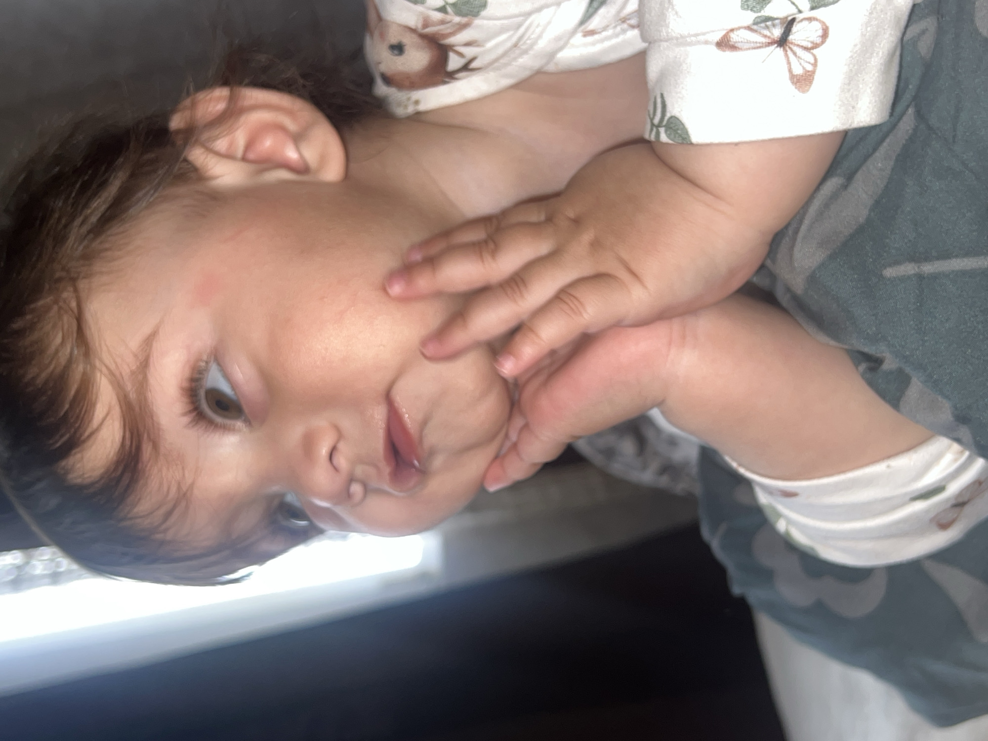 This close-up portrait, taken in Elena, Bulgaria, features a baby or young toddler. The child is depicted lying down, with its head tilted sideways, and its gaze directed towards the left side of the frame.
The baby has brown eyes and dark hair, with fair skin and full cheeks. Its mouth is slightly agape, conveying a relaxed or perhaps pensive expression. One small hand is gently placed against its cheek. A faint, small reddish mark is visible on the forehead near the hairline.
The child is dressed in a white long-sleeved top patterned with small brown deer, butterflies, and green leaves. Below the top, a dark green fabric with a subtle camouflage-like design is visible, likely a blanket or part of the child's clothing. The background is mostly dark and blurry, with a prominent bright, horizontal light source appearing along the bottom edge of the frame if the image were upright, suggesting an indoor setting with artificial light or a window. There is no visible text in the image.
