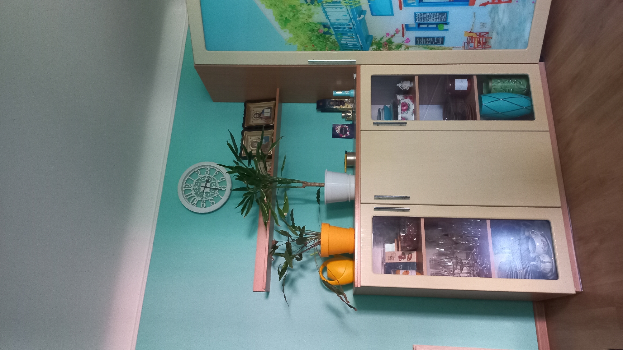 The image depicts a room interior with a light teal wall. On the wall hangs a white decorative clock. To the right of the clock, a cabinet with glass doors and a decorative picture on top is mounted on the wall. Two potted plants are visible: one in a white pot and another in an orange pot. A yellow watering can is also present, positioned next to the orange pot. The upper shelves of the cabinet display decorative objects, while the lower shelves contain glassware. The scene appears to be indoors. There is no information in this image that identifies it being in Kremenchuk, Ukraine.