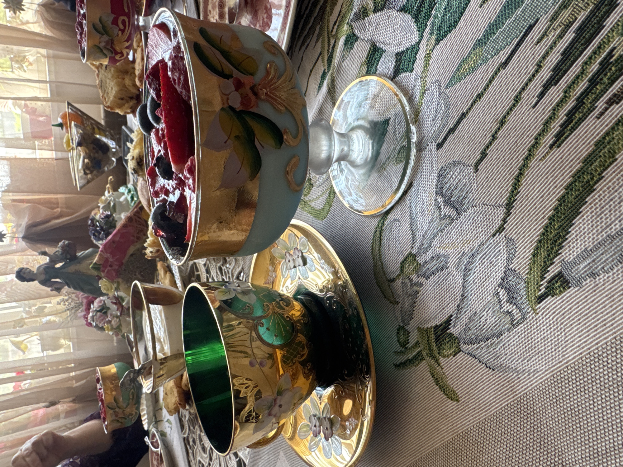 The image presents a tabletop scene, possibly in a residential setting in Plovdiv, Bulgaria. The focus is on a collection of ornate glassware, including what appear to be dessert bowls and cups. One bowl contains a fruit dessert, possibly berries and other soft fruits. The glassware features intricate designs and gold accents, suggesting a vintage or antique aesthetic. The tablecloth has a textured pattern with floral elements. The backdrop includes a window with sheer curtains and hints of other decorative items, such as figurines and flowers, indicating a decorated interior space. A person's hand is visible in the lower-left corner.