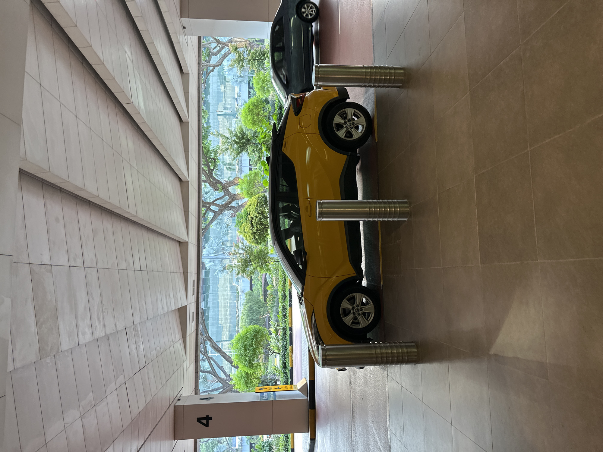 A yellow car is parked in a covered area with a tiled floor and ceiling. The car is a hatchback with a black roof and tinted windows. It is surrounded by metal bollards. In the background, there is a row of trees and a building with a glass facade. A dark-colored sedan is also visible in the background. A sign on a pillar near the yellow car has the number "4" on it. This scene is likely in Singapore, given the lush greenery and modern architecture.