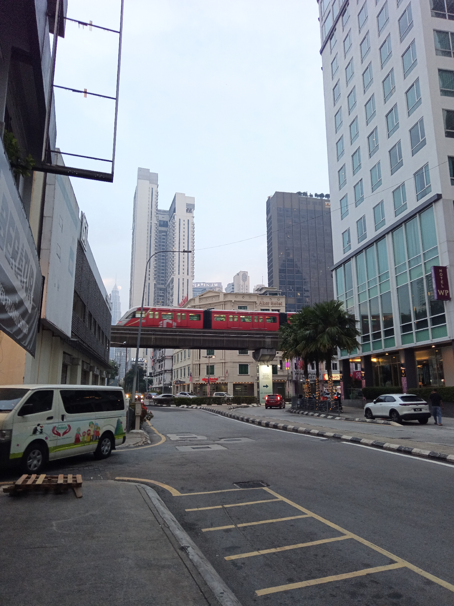 A red monorail train travels on an elevated track above a street in Kuala Lumpur, Malaysia. The train is passing in front of several tall buildings, including a modern white high-rise with a repeating window pattern on the right and a dark, rectangular skyscraper in the background. The street below is paved and has yellow and white markings. A white van with colorful graphics is parked on the left side of the street. A white car is parked on the right side of the street, and a person is standing near it. Trees are visible between the buildings and along the street. The sky is overcast.