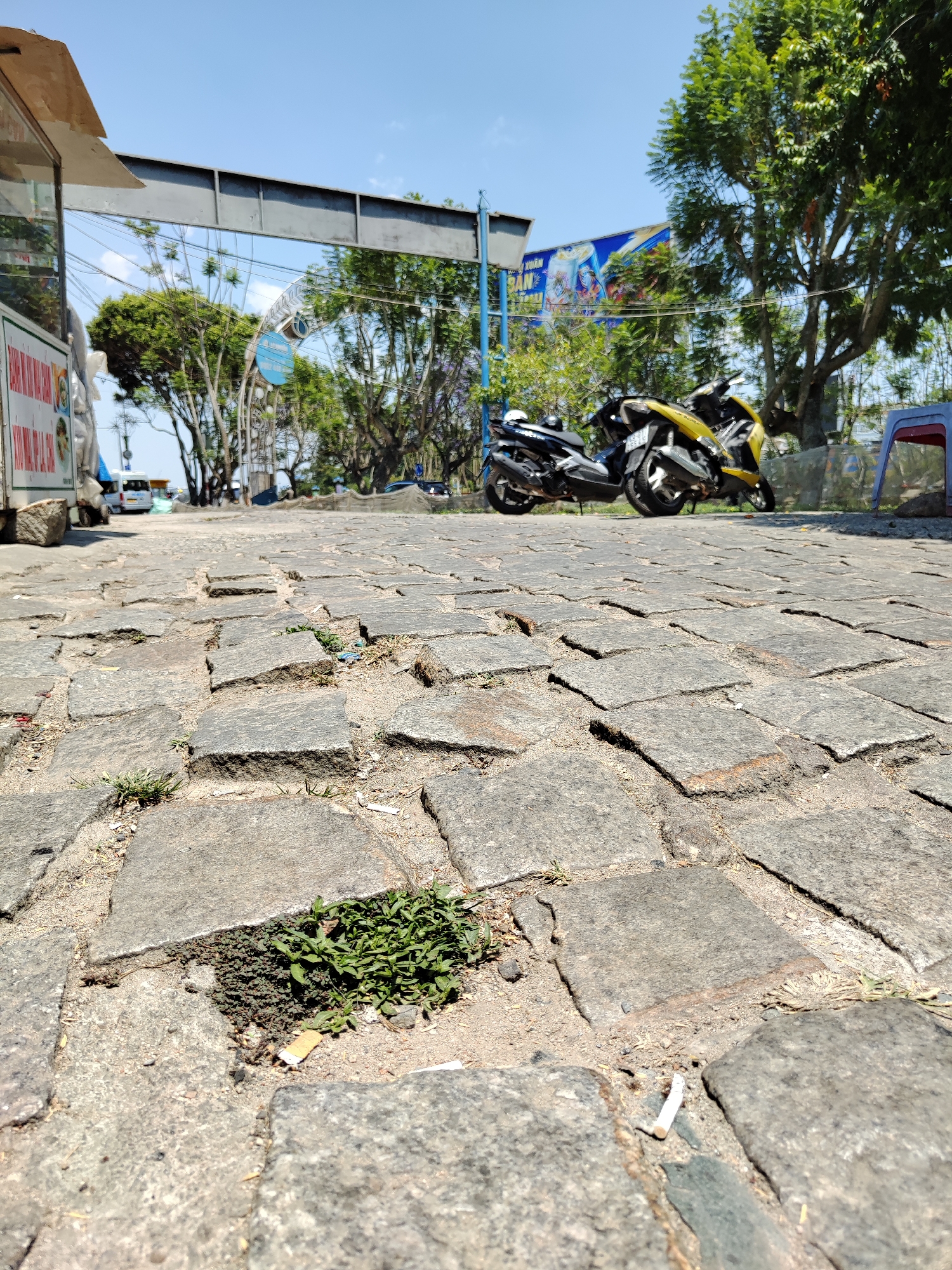 The image shows a stone-paved road in Da Lat, Vietnam. Weeds grow in the cracks between the stones. In the background, two parked motorbikes, one black and one yellow, sit near a sidewalk. Beyond the bikes are trees, a sign, and building structures. A blue sky is visible above. The photo is taken from a low angle, focusing on the stonework and weeds in the foreground. A cigarette butt lies on the ground amongst the stones.