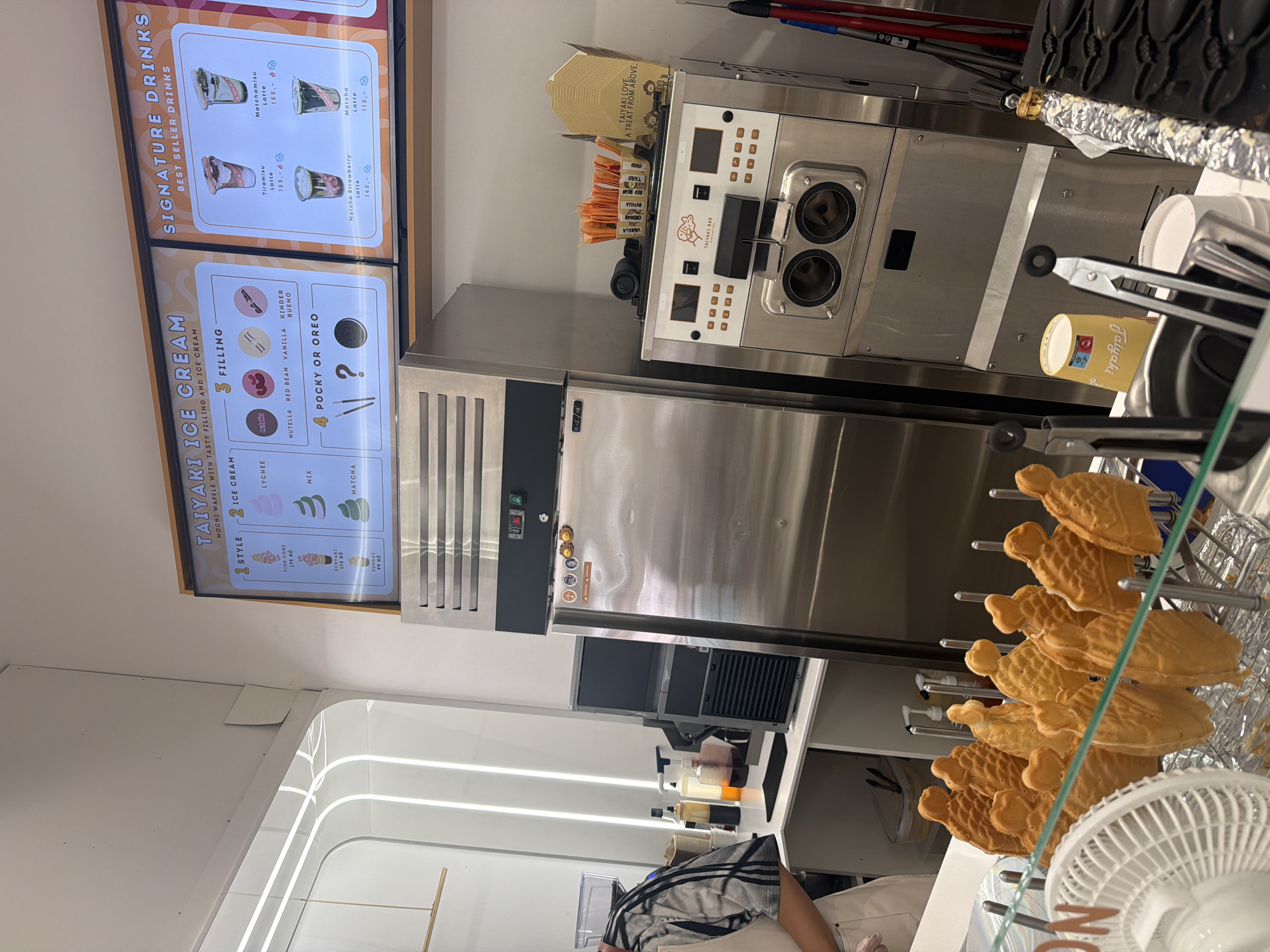 The image depicts the interior of an ice cream shop. A large, stainless steel commercial refrigerator is the central focus, with a digital display showing "36". To the right of the refrigerator is an ice cream machine with two dispensing nozzles. Above the refrigerator, a digital menu board displays "TAIYAKI ICE CREAM" and "SIGNATURE DRINKS". The menu lists various ice cream styles, fillings, and drinks with prices, suggesting the location is in Prague, Czechia. In the foreground, several fish-shaped taiyaki pastries are displayed on a metal rack. A white fan is positioned to the left. The background shows shelves with bottles, cups, and other shop supplies. A person wearing a patterned shirt and apron is partially visible on the left, interacting with the counter area.