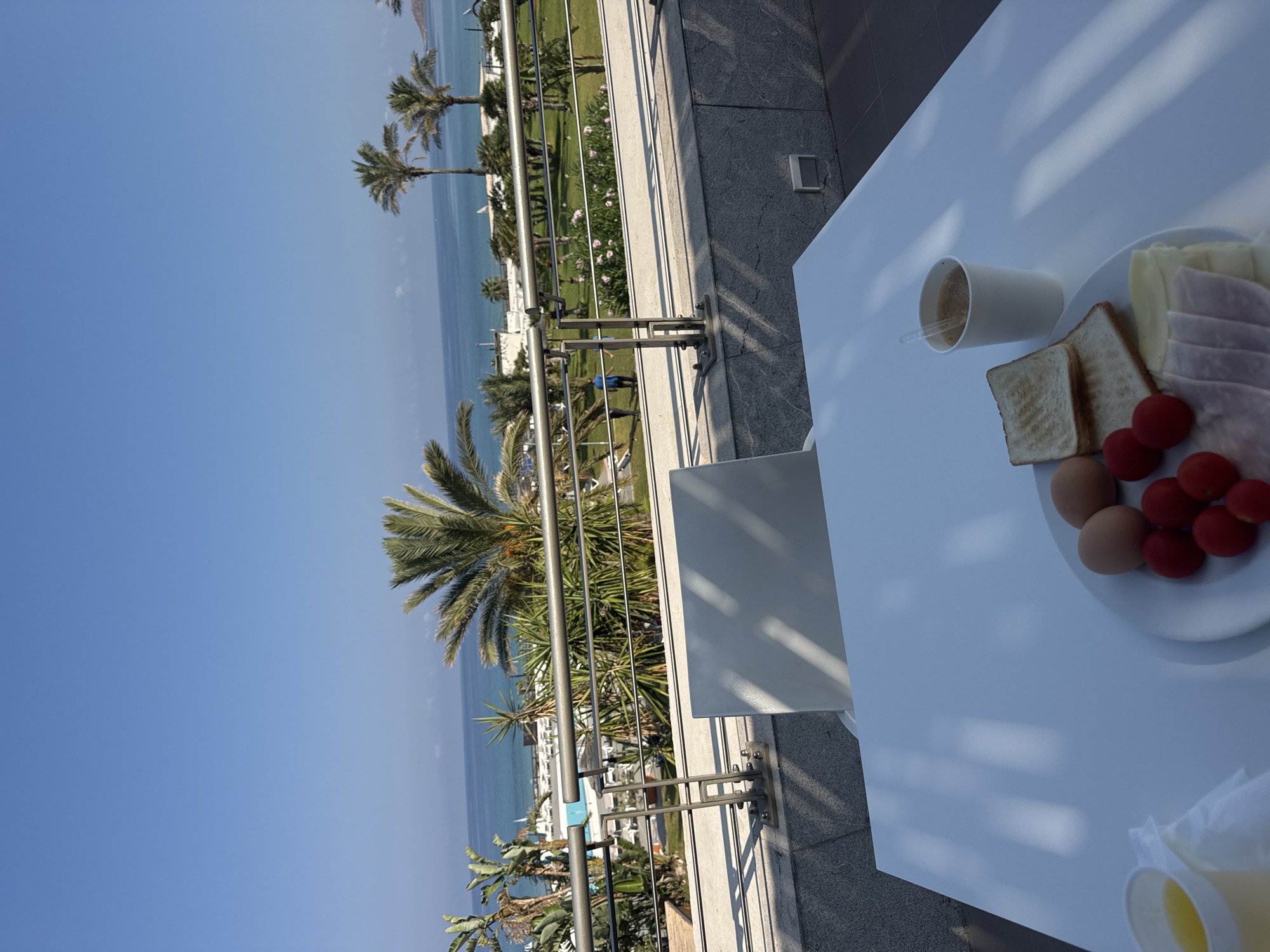The image shows a breakfast spread on a white table with a view of the Mediterranean Sea and palm trees in Isola delle Femmine, Italy. On the table, there is a plate with two boiled eggs, cherry tomatoes, two slices of toasted bread, sliced ham, and sliced cheese. A white paper cup containing a beverage, possibly coffee or tea, and a cup of orange juice are also on the table. A balcony railing with a metal bar runs horizontally across the middle ground, separating the foreground table from the scenic background. Beyond the railing, a well-maintained lawn with scattered palm trees and some flowering bushes is visible. In the far background, the turquoise sea meets the clear blue sky, with a hint of the coastline on the horizon. A swimming pool and some lounge chairs are also visible near the water. The scene is bathed in bright sunlight, casting shadows on the table and balcony. There are no people in the immediate foreground.