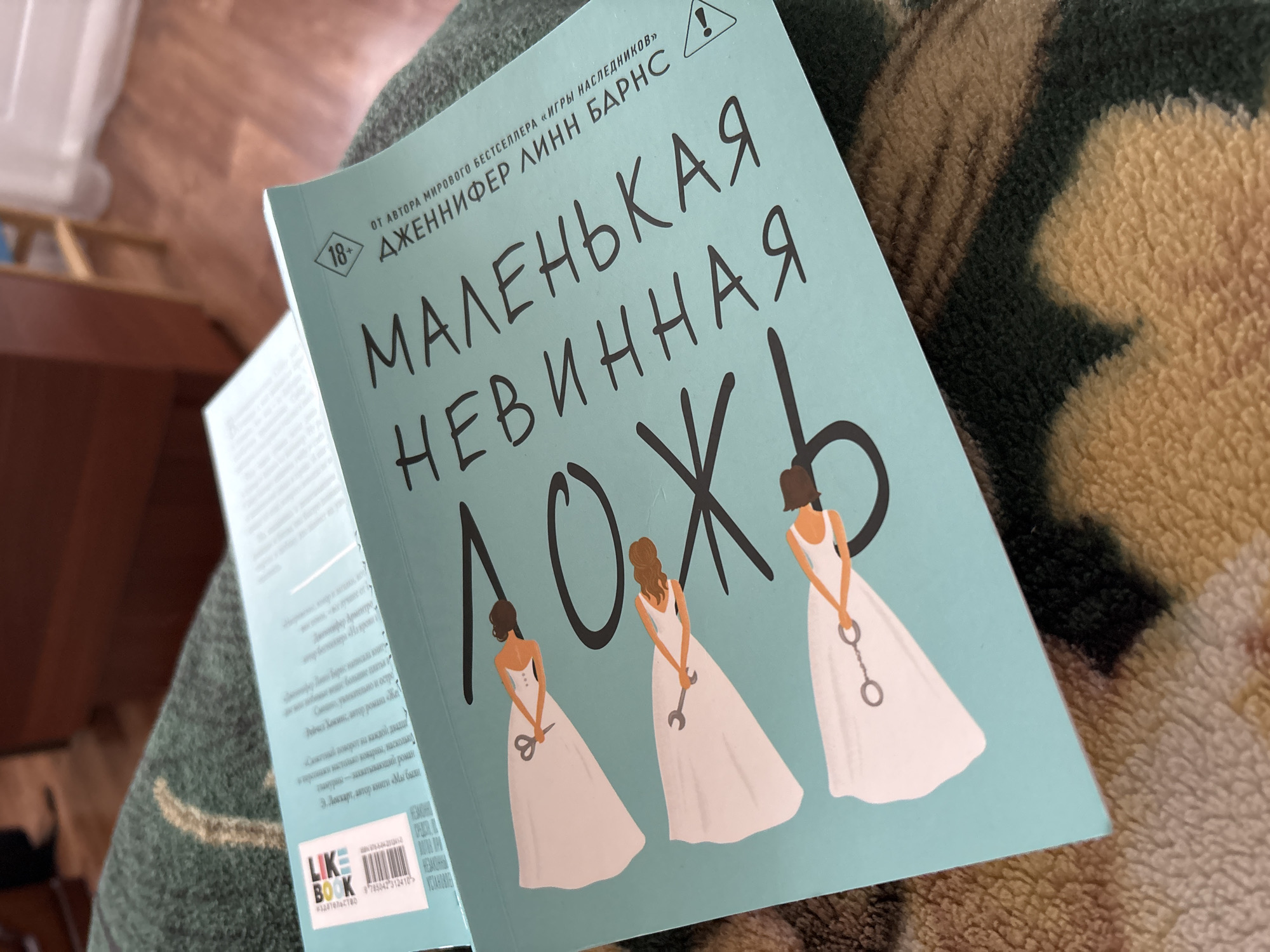 A close-up shot focuses on a book titled "Маленькая невинная ложь" (Little Innocent Lie) by Jennifer Lynn Barnes. The book's cover is light blue and features three stylized women in white dresses, each holding a different tool: scissors, a wrench, and handcuffs. The book appears to be in Russian, as indicated by the title and text on the cover.
The book is resting on a patterned surface. A portion of the book's spine is visible, displaying additional text and a logo labeled "LIKE BOOK издательство" (LIKE BOOK Publishing). The image has good lighting and contrast. The surroundings are slightly blurred, indicating a shallow depth of field. The city of Izhevsk and Russia in general cannot be determined from the image.