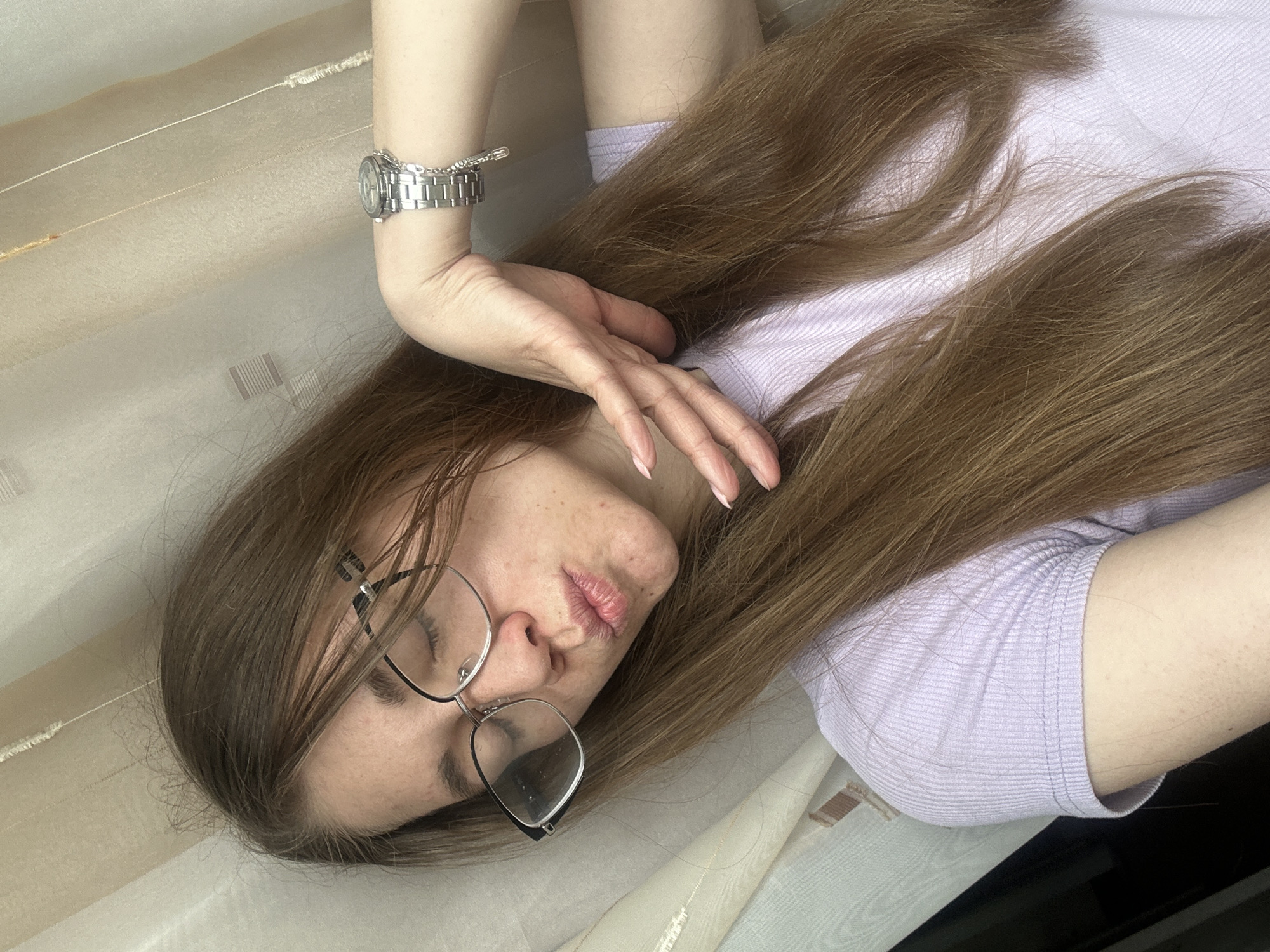 A young, fair-skinned woman with long, light brown hair is lying down. She is wearing eyeglasses and a light purple ribbed shirt. A silver-colored watch is on her left wrist. Her left hand is positioned near her face, with her fingers slightly curled. The background appears to be a cream and light brown striped surface, possibly a bed or sofa. There is no verifiable information present to definitively confirm that this image was taken in Chaikovsky, Russia.