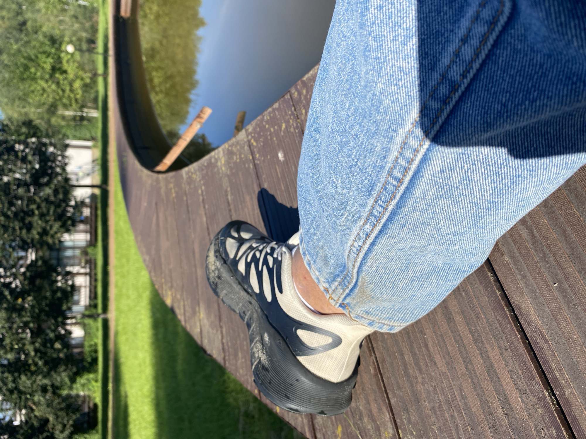 A first-person perspective shot shows a person's leg clad in blue jeans and a black and beige sneaker resting on a wooden boardwalk. The boardwalk curves around a body of water, which appears to be a pond or a shallow lake. The water reflects the surrounding trees and sky. Beyond the boardwalk and water, a well-maintained lawn stretches towards a row of buildings, likely apartments or houses, bordered by trees. The scene is illuminated by bright sunlight, casting shadows on the boardwalk. The location is identified as Chulis, Russia.