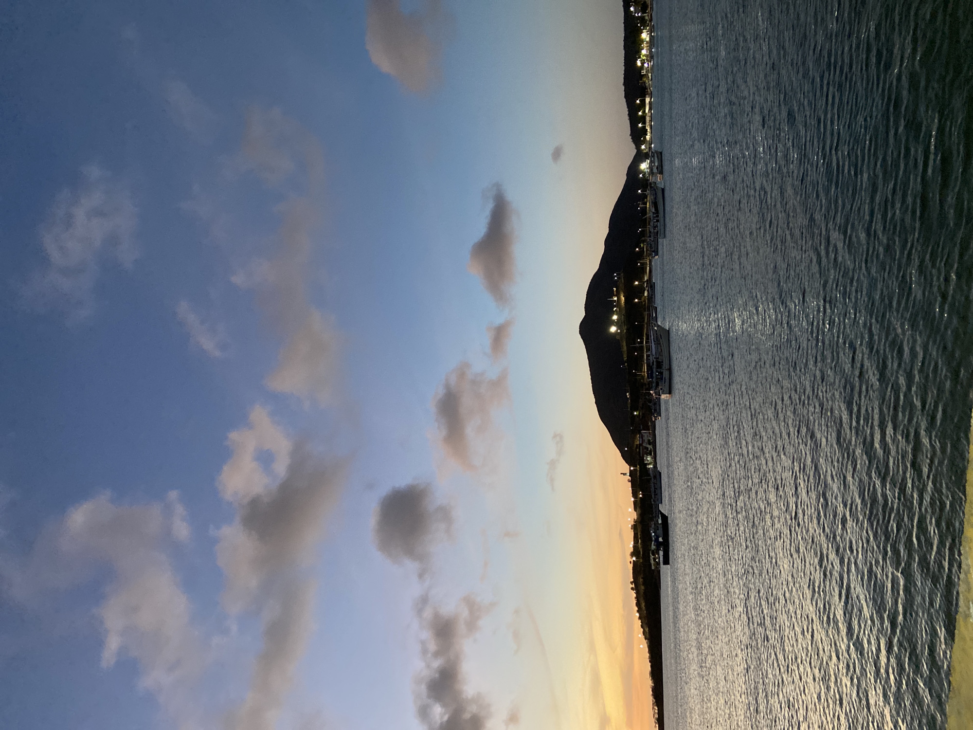 The image displays a serene coastal landscape captured during dusk or early twilight, set against the backdrop of Juan Griego, Venezuela. The sky dominates the upper portion of the frame, showcasing a beautiful gradient from a soft blue at the zenith to warmer tones of yellow and orange nearer the horizon, indicating the setting sun. Scattered cumulus clouds, mostly greyish-white, punctuate the blue expanse.
Below the colorful sky, a calm body of water stretches across the mid-ground, its surface gently rippled and reflecting the fading light. Along the distant horizon, a dark silhouette of a mountainous or hilly landmass is visible. Numerous bright artificial lights twinkle along the base of this landmass, suggesting the presence of a coastal town, a pier, or a marina. Indistinct shapes that could be boats or docks are subtly visible near these lights.
No people or specific activities are discernible in the tranquil scene. A blurry, yellowish vertical element occupies the far right edge of the foreground, hinting that the photograph might have been taken from a structure like a railing or a boat. The overall atmosphere conveyed is one of peace and natural beauty as daylight fades over the coastal environment. There is no visible text in the image.