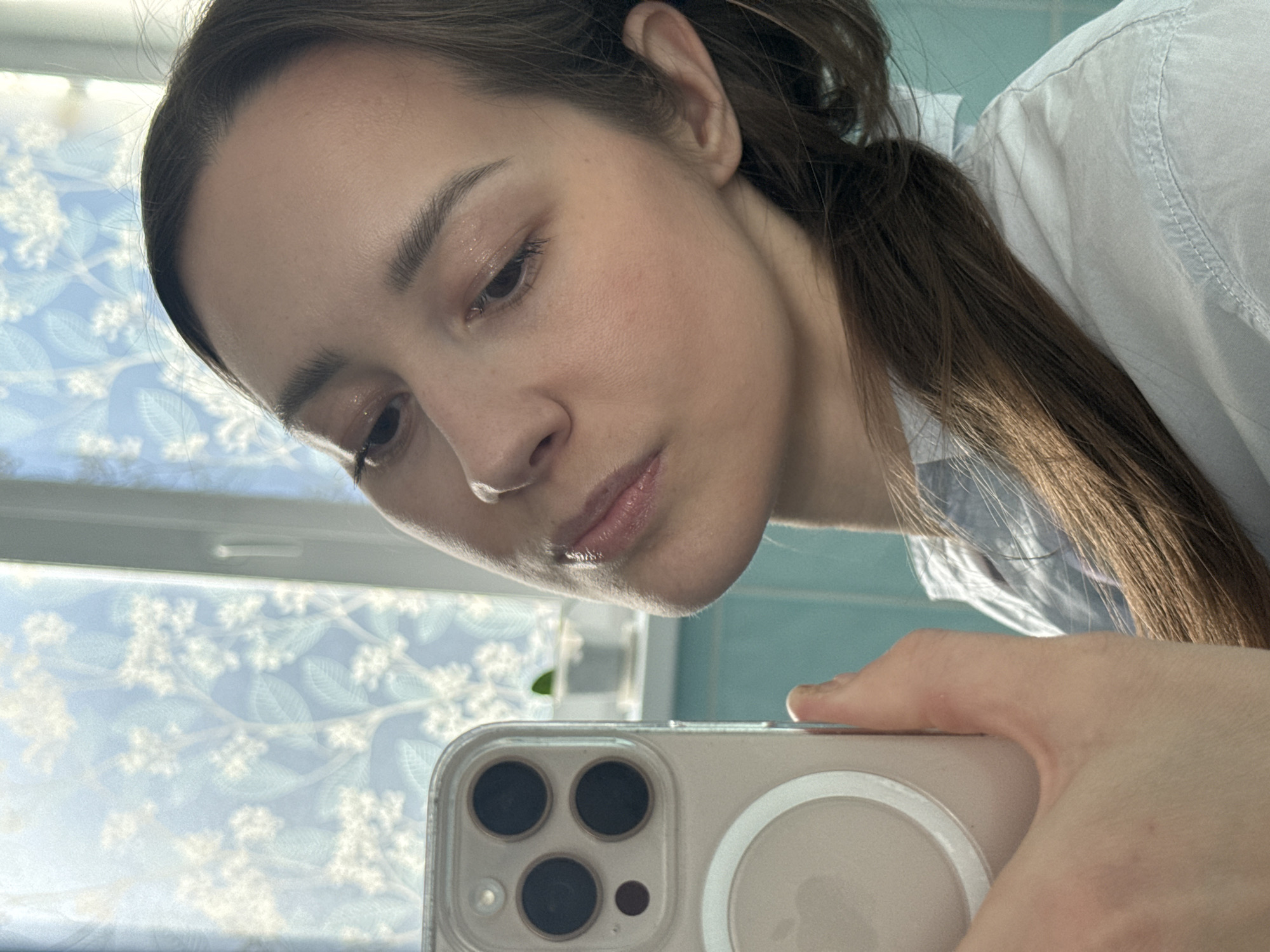 The image displays an angled, close-up selfie of a female individual taken indoors in the city of Chaikovsky, Russia. The subject has long brown hair, wears a light-colored collared shirt, and has makeup, including glitter eyeshadow and lip gloss. She holds an iPhone, identifiable as a Pro Max model in a clear MagSafe case, with her right hand in the lower left quadrant of the frame. Her gaze is directed downwards. The background features a window covered with a translucent decorative film displaying a pattern of white flowers and light green leaves against a light blue background, admitting natural light. A portion of a turquoise wall or tiled surface is visible to the right, along with a small green potted plant on a ledge near the window.